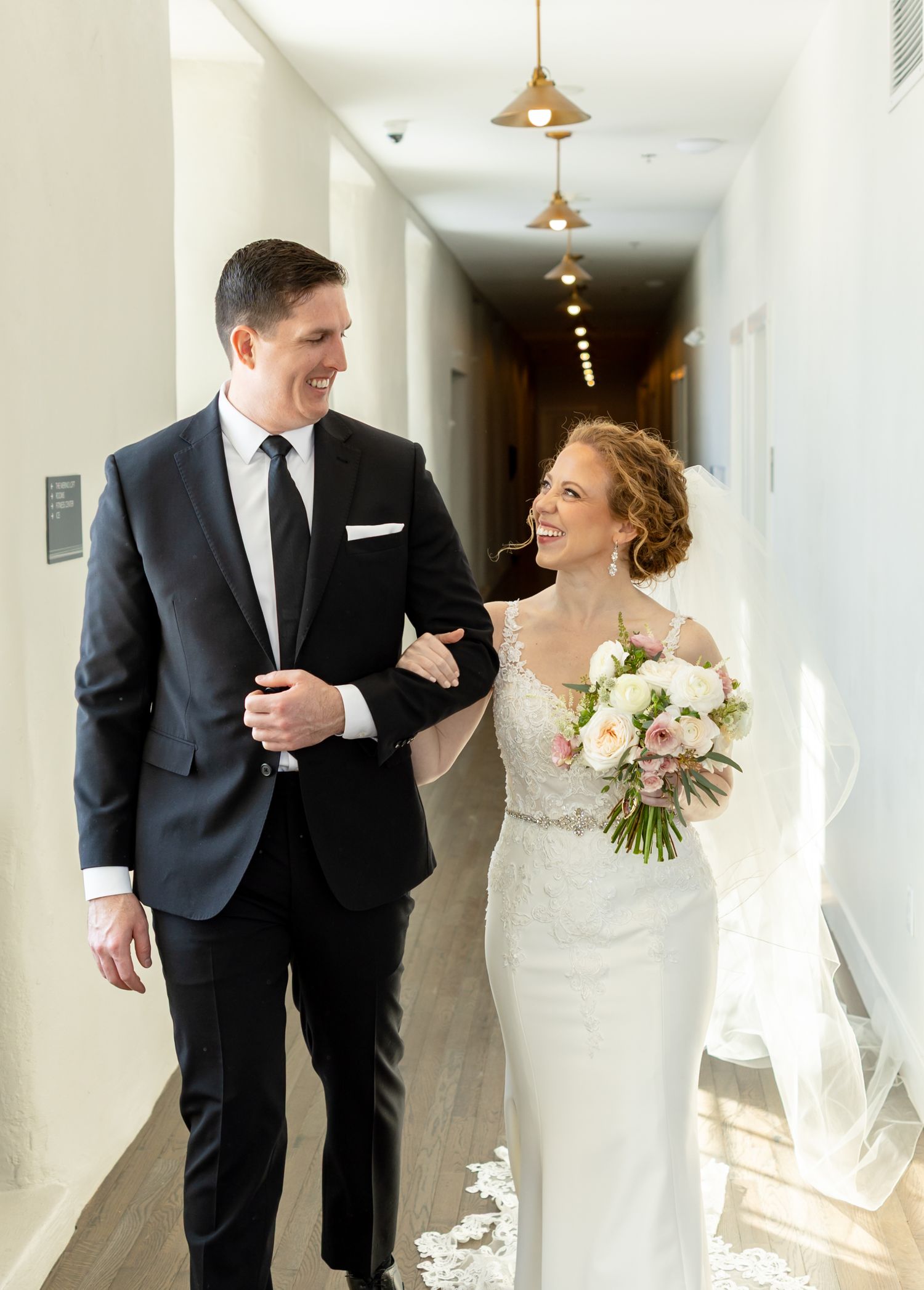 Happy couple walking down bright hallway with pendant lights and white bouquet.