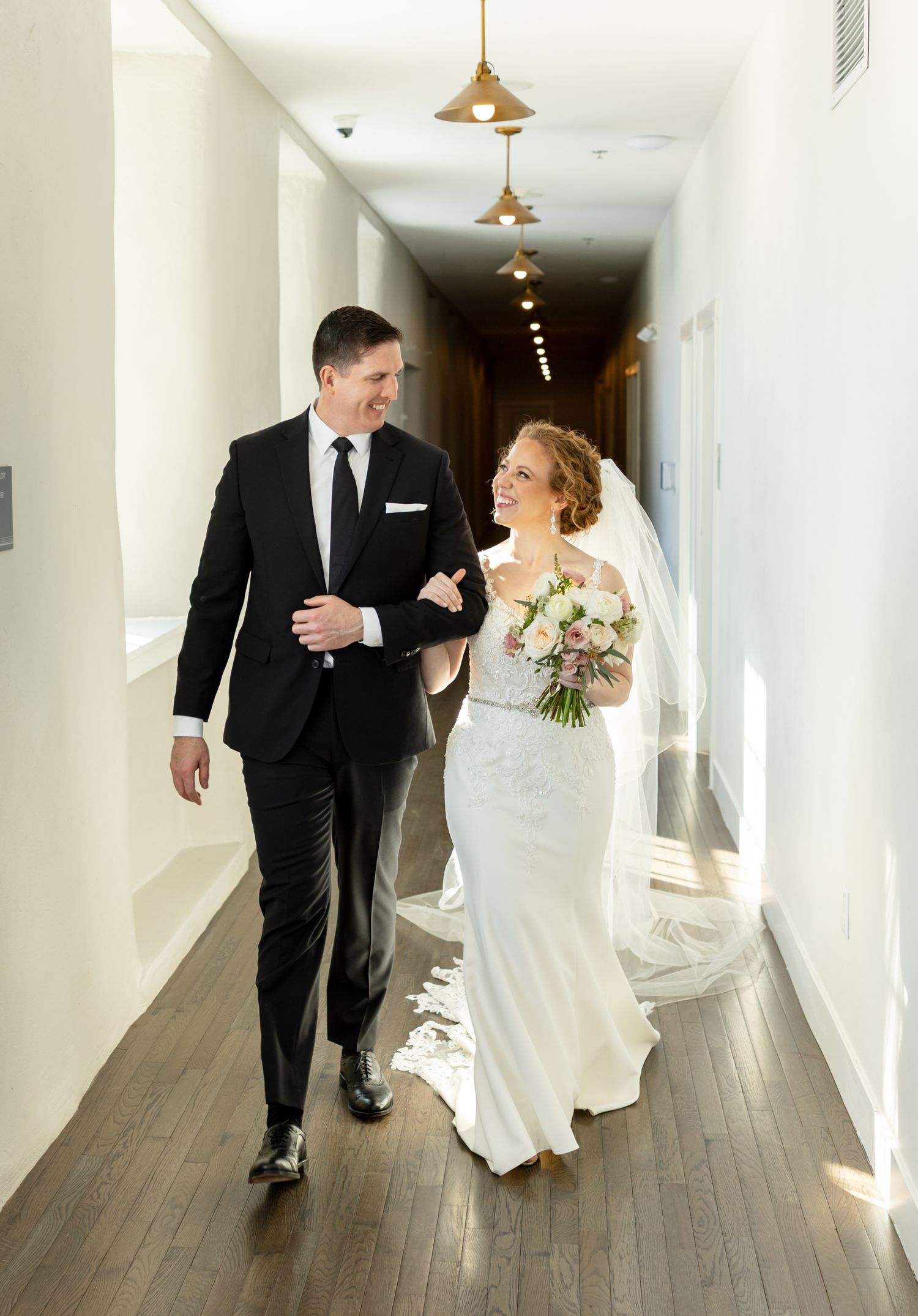A bride and groom share a joyful moment walking down a bright hallway with hardwood floors and pendant lighting.