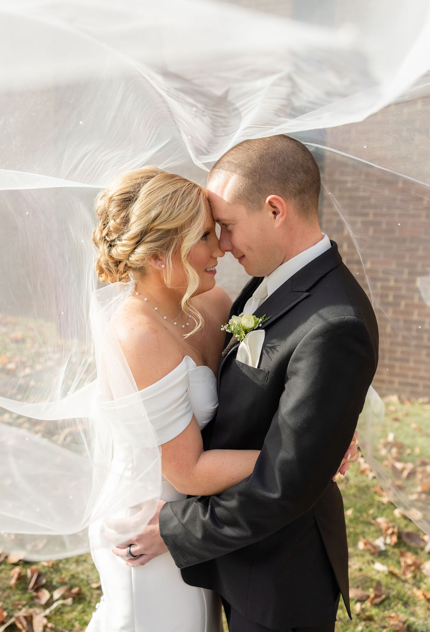 A romantic wedding moment captured under a flowing veil with autumn leaves in the background.