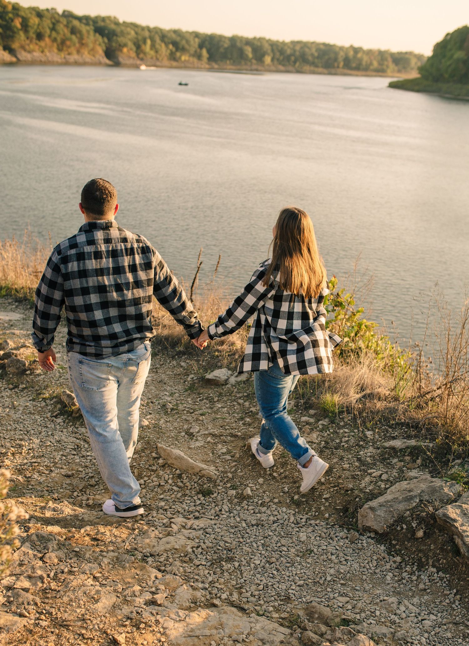Two people in matching plaid shirts walk along a rocky lakeside path at sunset.