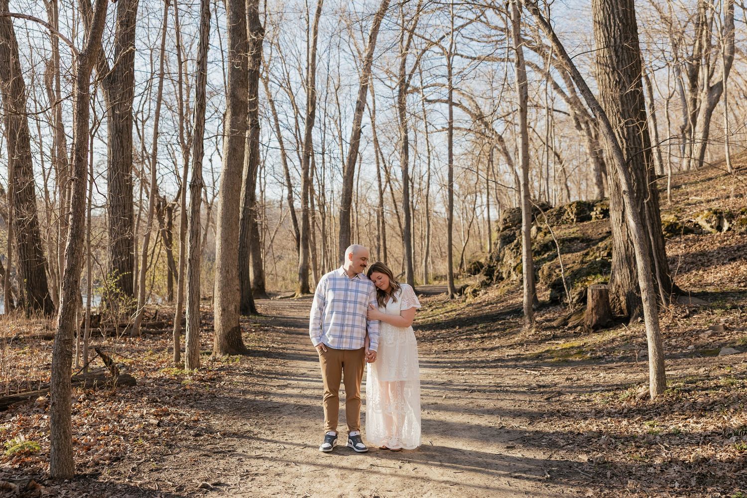 Couple embracing on a wooded forest path surrounded by bare trees in a romantic outdoor engagement photo.