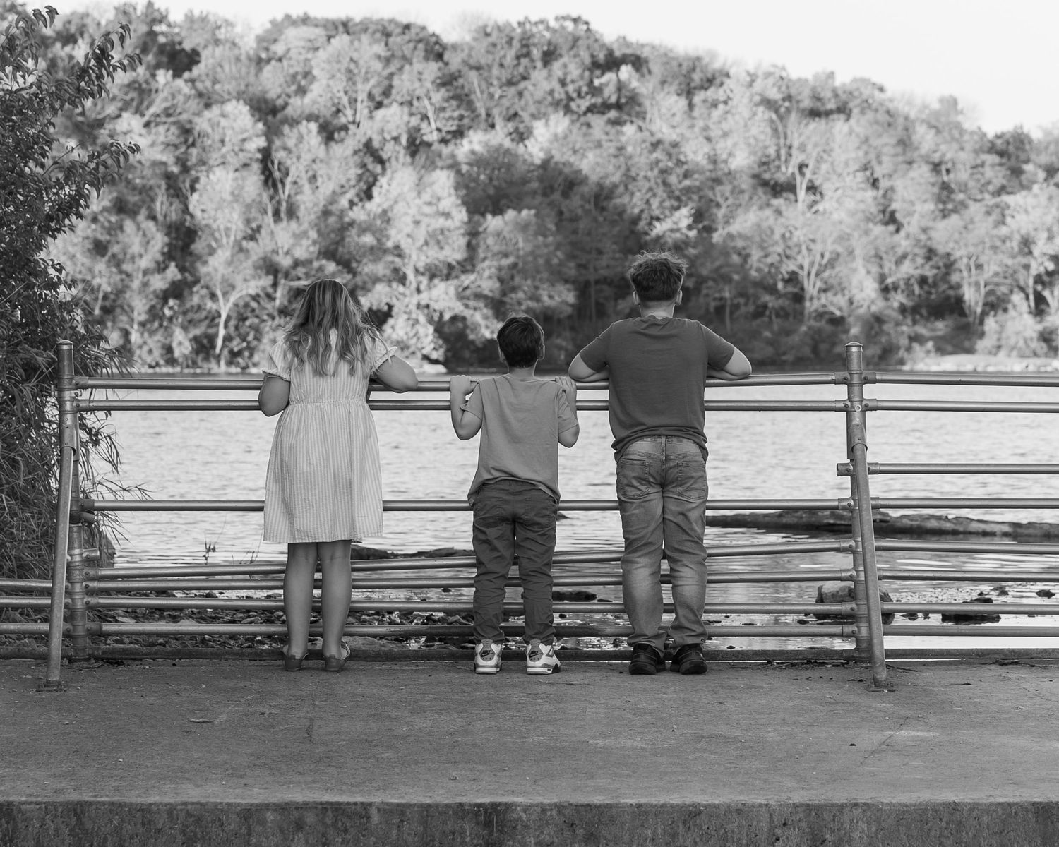 Three children stand at a railing overlooking a river surrounded by lush trees in a black and white photo.