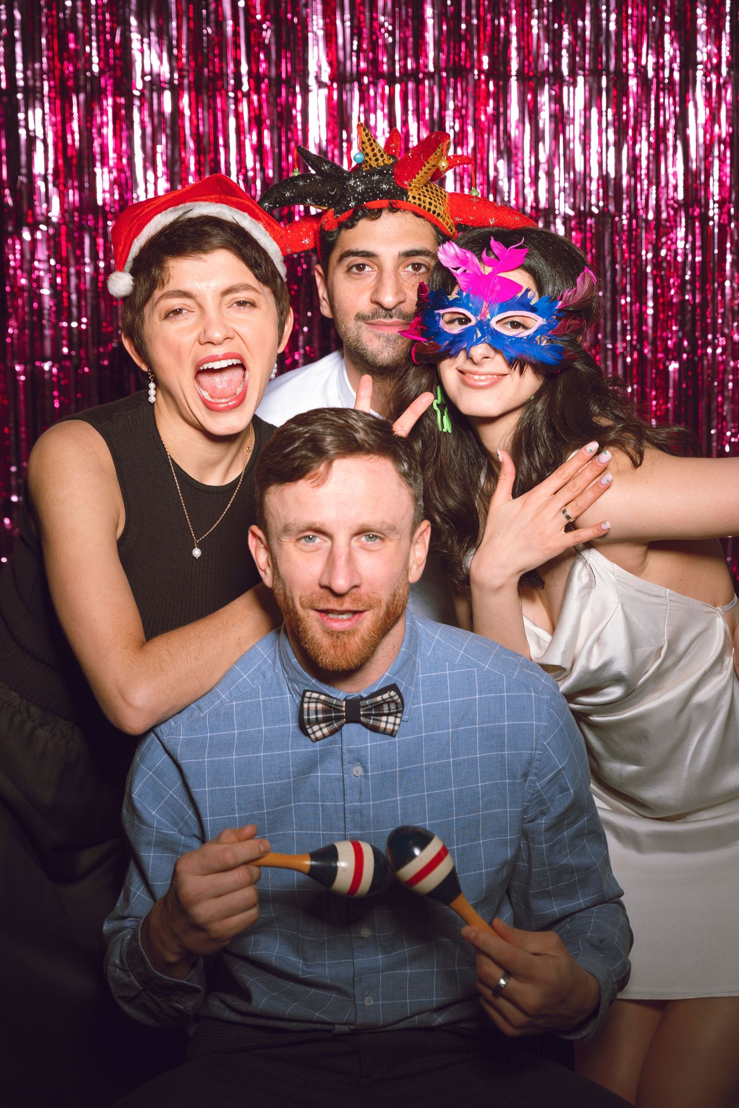Party guests pose with props and masks against pink sparkly backdrop.
