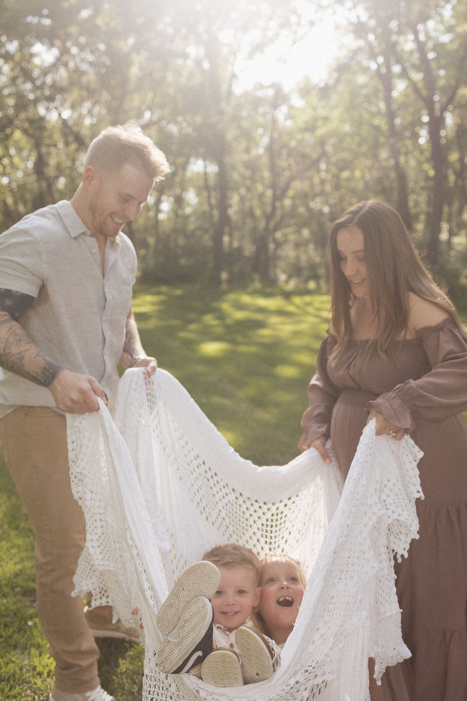 family of four swinging their two children in a white woven blanket playfully in an open green space surrounded by tall green trees
