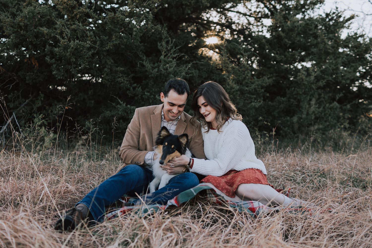 couple sitting together on blanket in tall grasses with pine trees while petting their dog at lake olathe