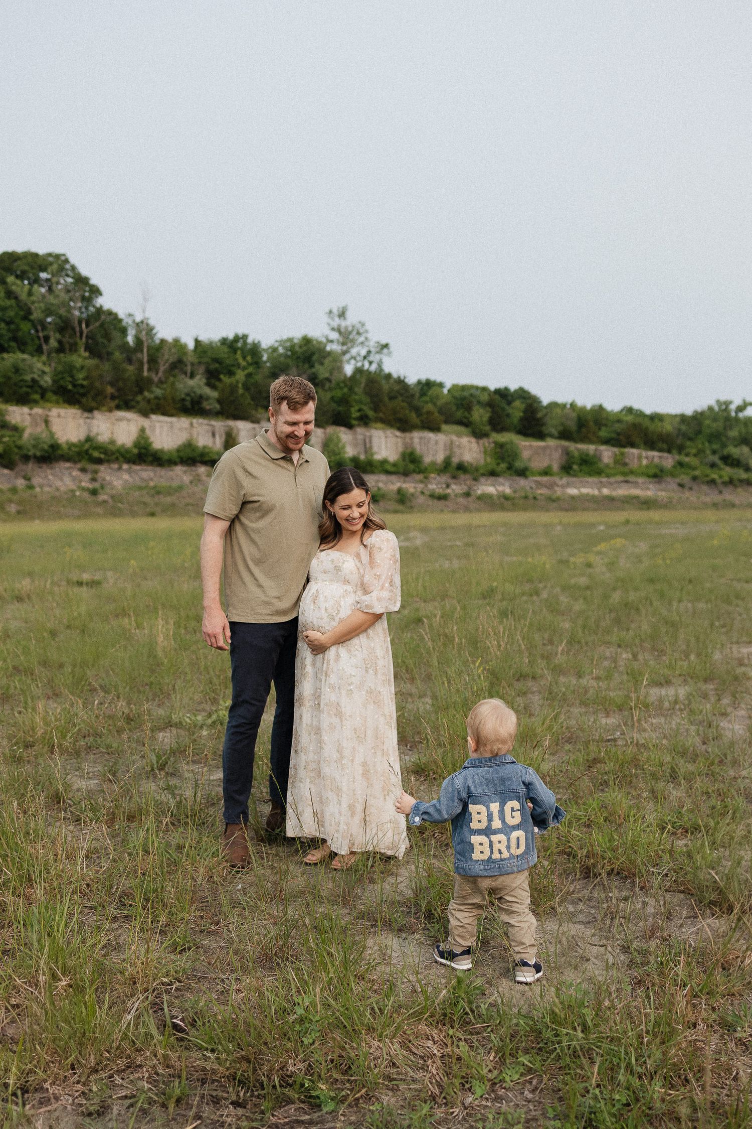 husband and wife smiling down at toddler son with big bro written on the back of his jean jacket with blue springs lake rock wall
