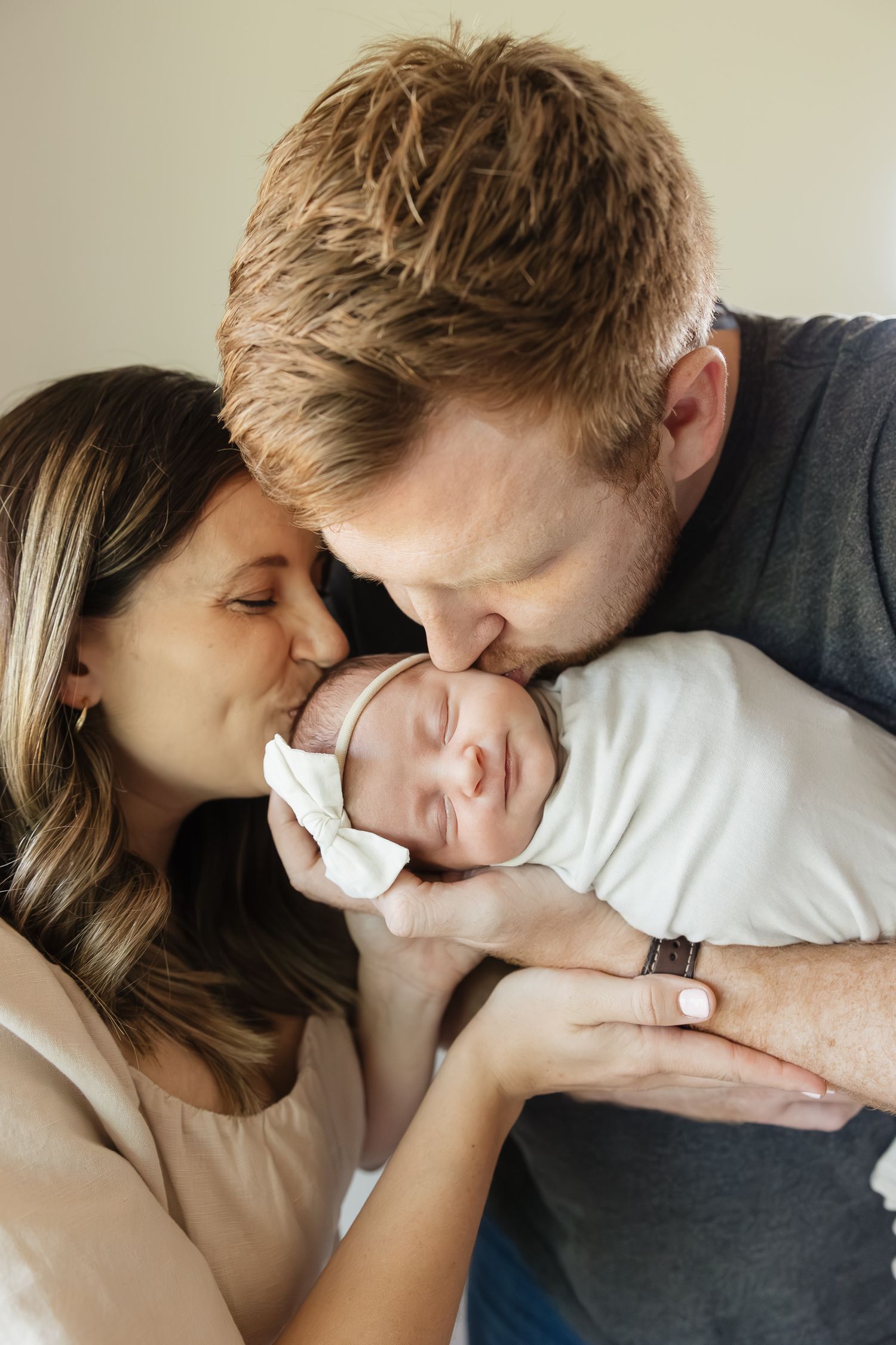 mom and dad kiss cheek and head of newborn baby girl smirking at camera while sleeping family photographer kansas city