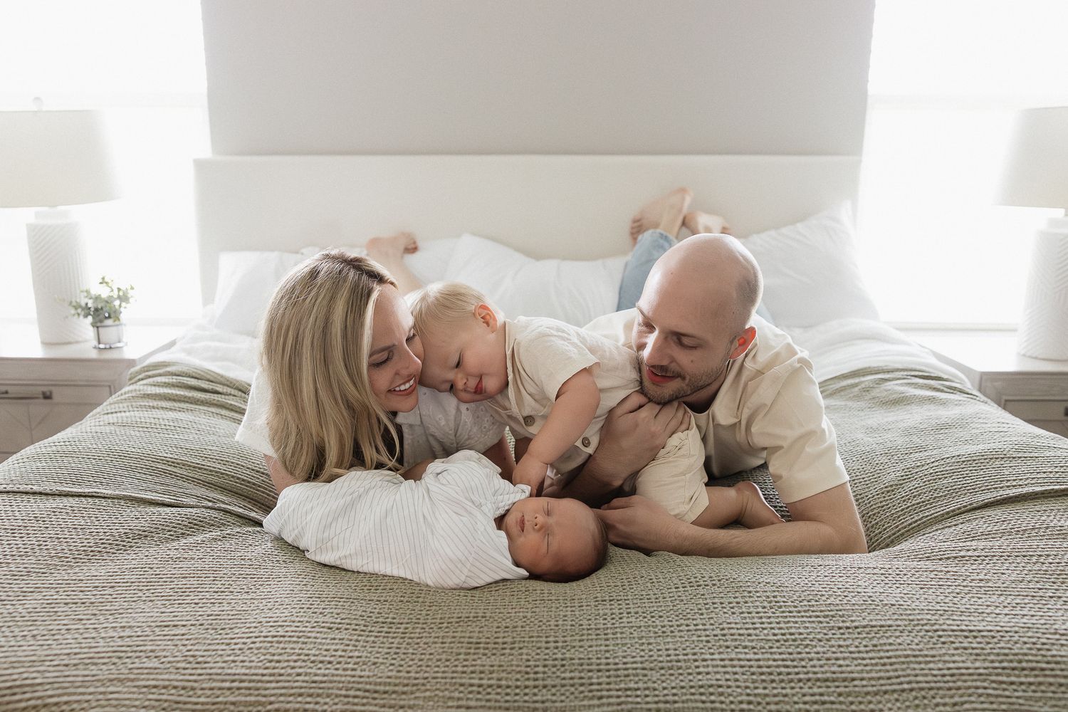 family laying on master bed on stomach looking at baby brother laying infront of them toddler touches baby's ear