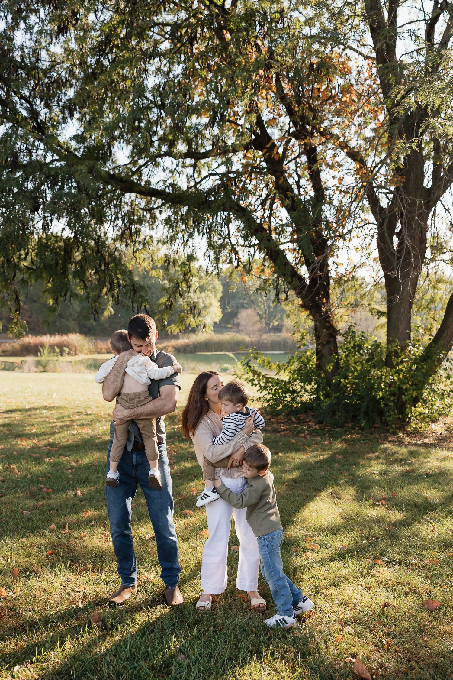 family of 5 hug and snuggle under a big tree with pond in the background in overland park family photographer kansas city
