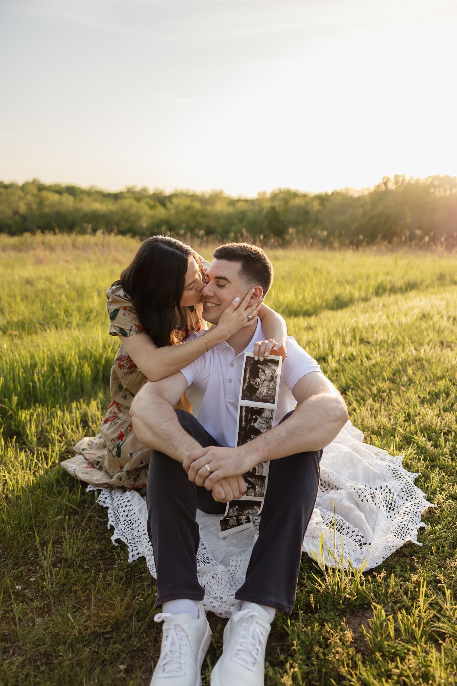 dad to be sitting on white blanket around tall prairie grasses with wife giving him a kiss on the cheek while holding a sonogram photo