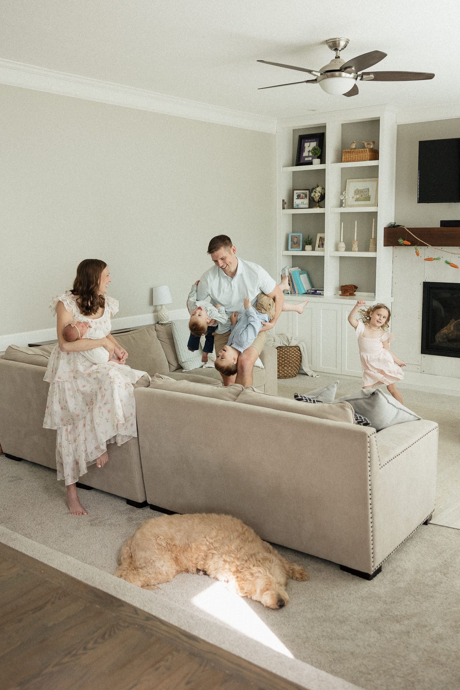family dancing together in living room dad wrestling two boys mom holding newborn baby daughter dance by dog laying on carpet
