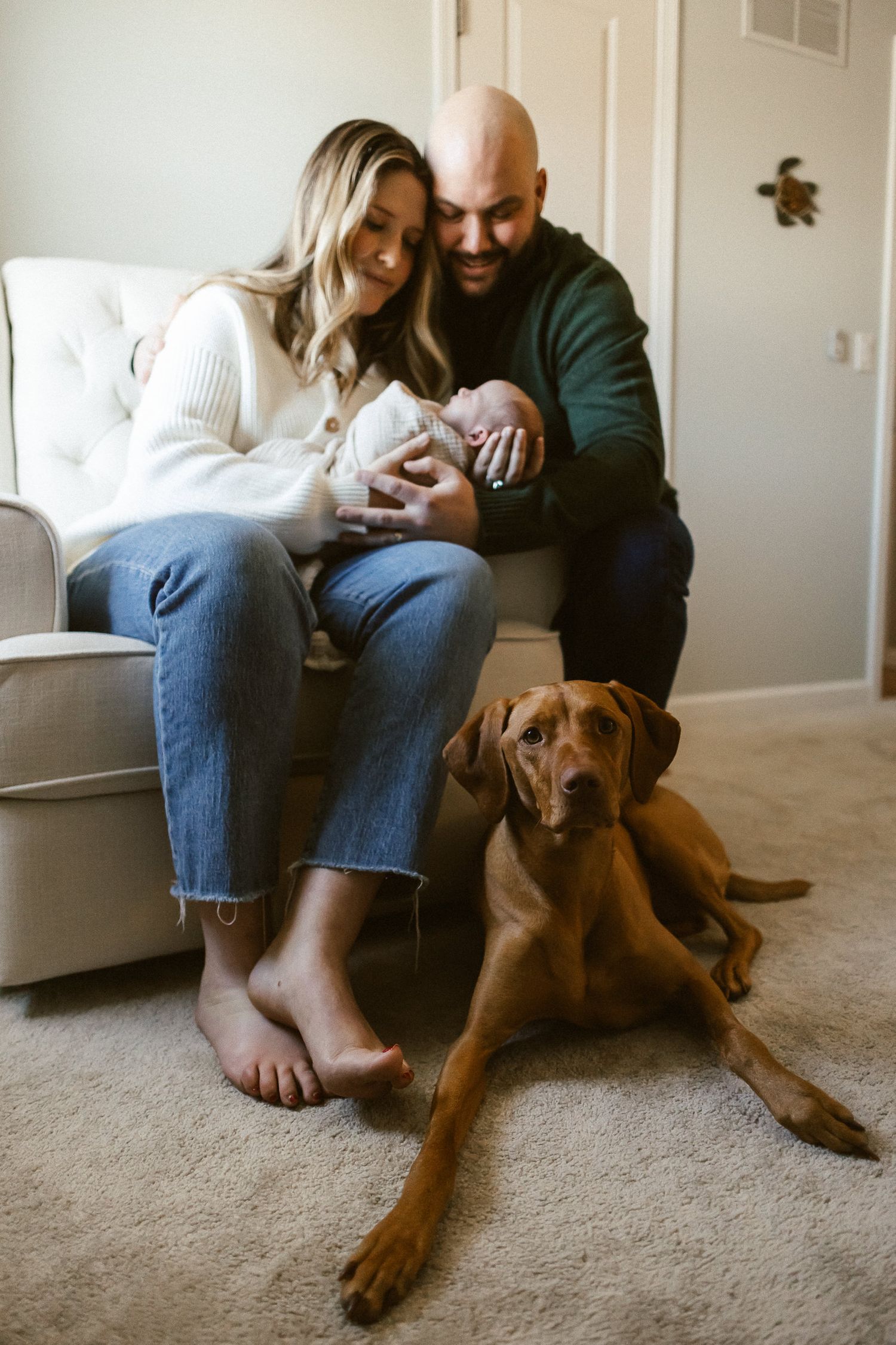 Mom and dad holding newborn baby girl while sitting in a rocking chair with their chocolate lab laying on the floor at their feet