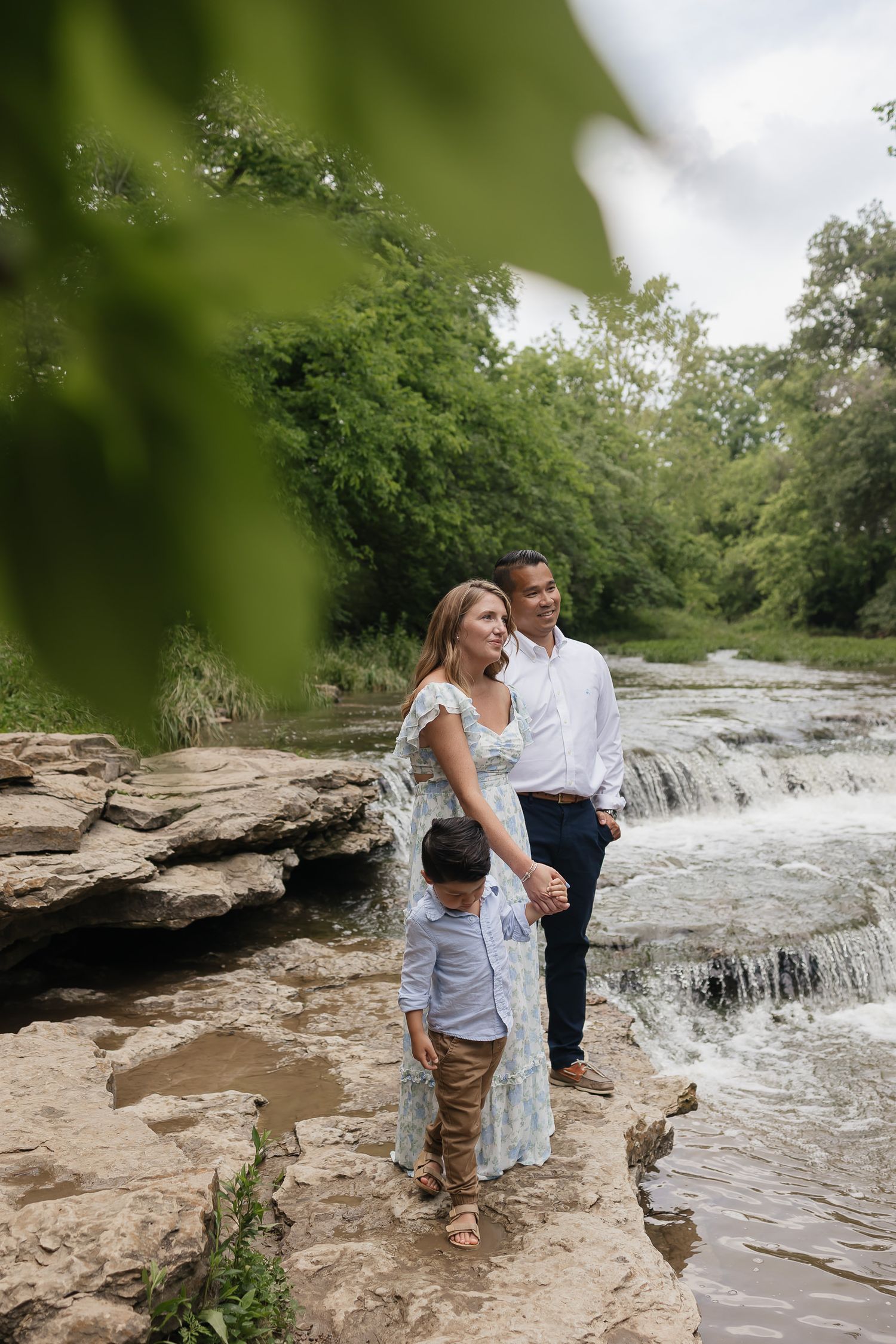 family of three stand by a rushing river staring at the clouds with green leaves in the foreground