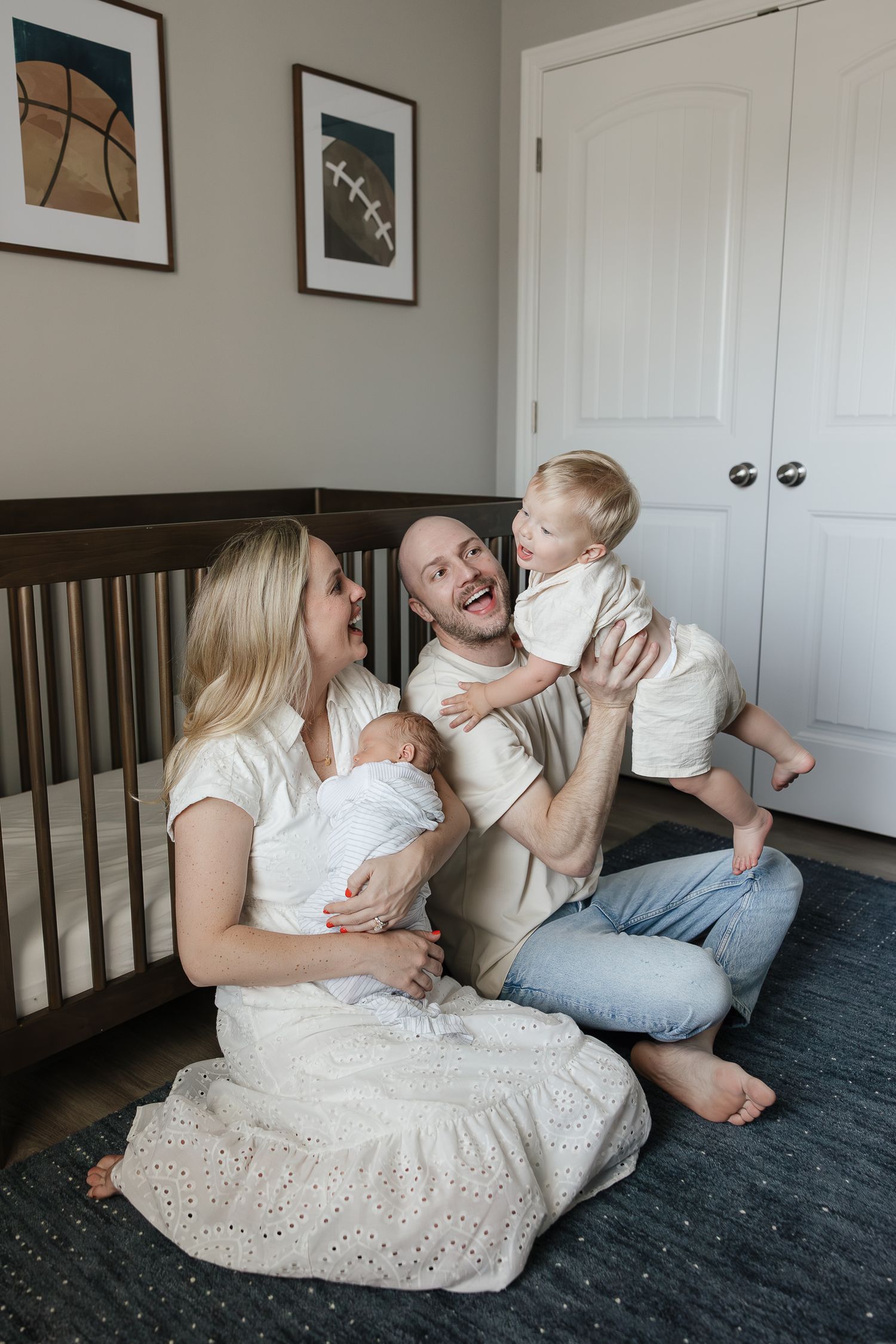 husband and wife sit on nursery floor holding newborn baby while playing with smiling toddler family photographer kansas city