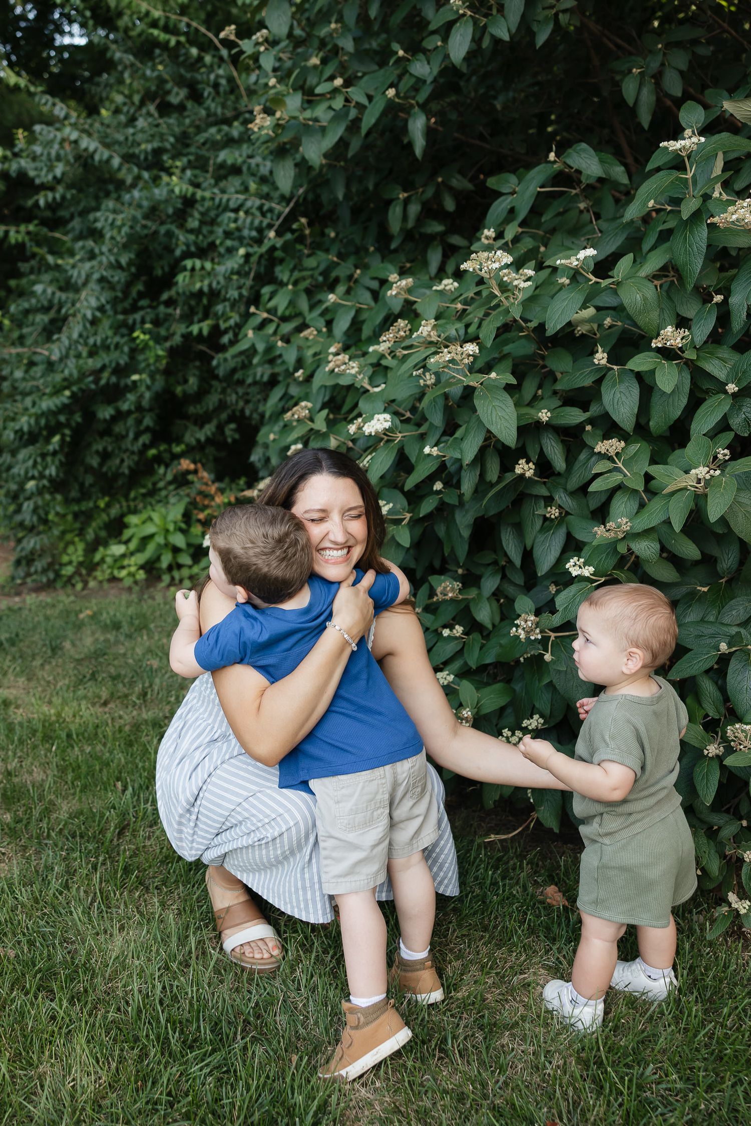 mom crouched down giving her toddler son a hug next to blooming bushes at antioch park photographer overland park ks
