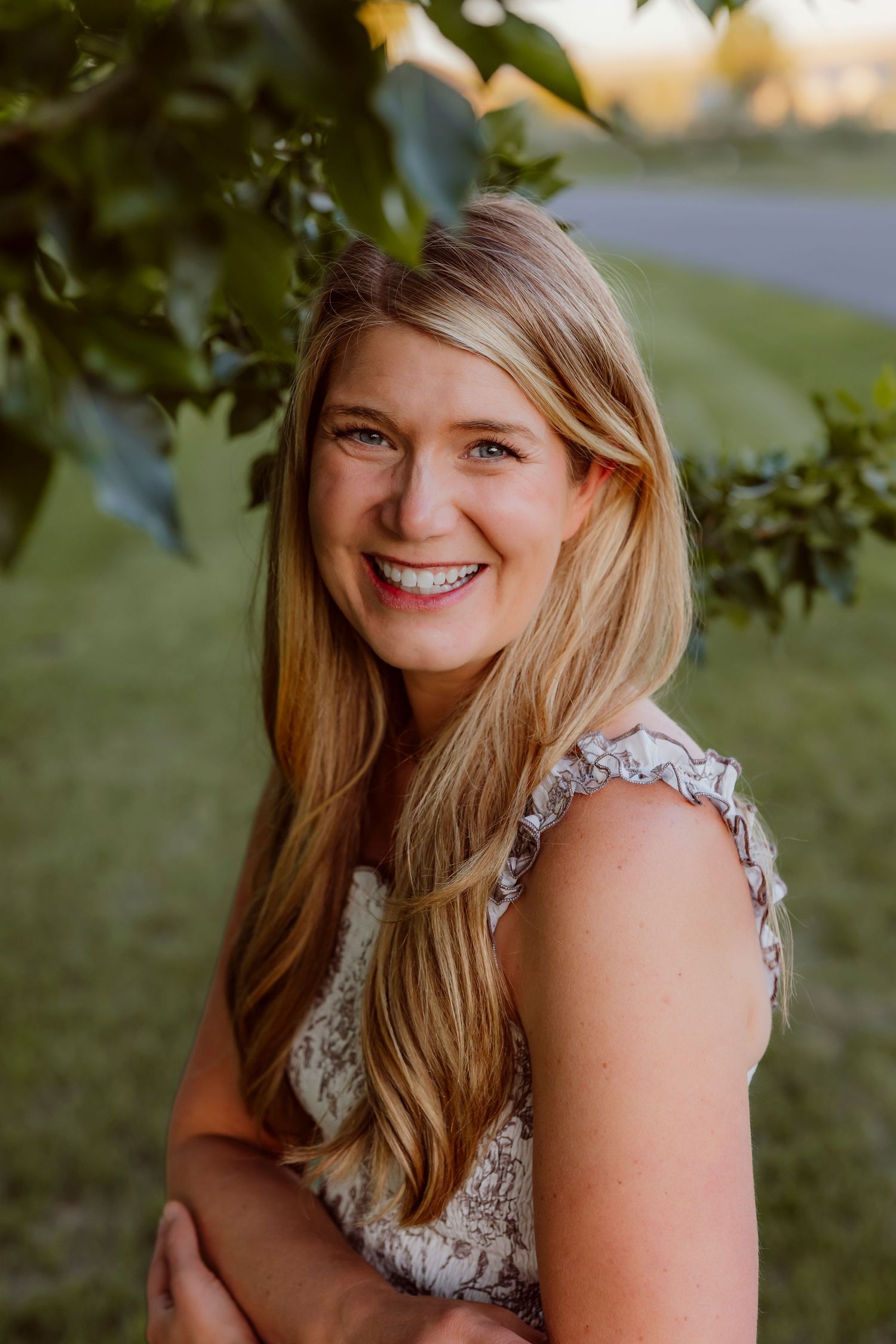 Smiling blonde woman in lace top standing under green tree branches in a sunny outdoor setting. Mackenzie Scheie Photographer Best in MT