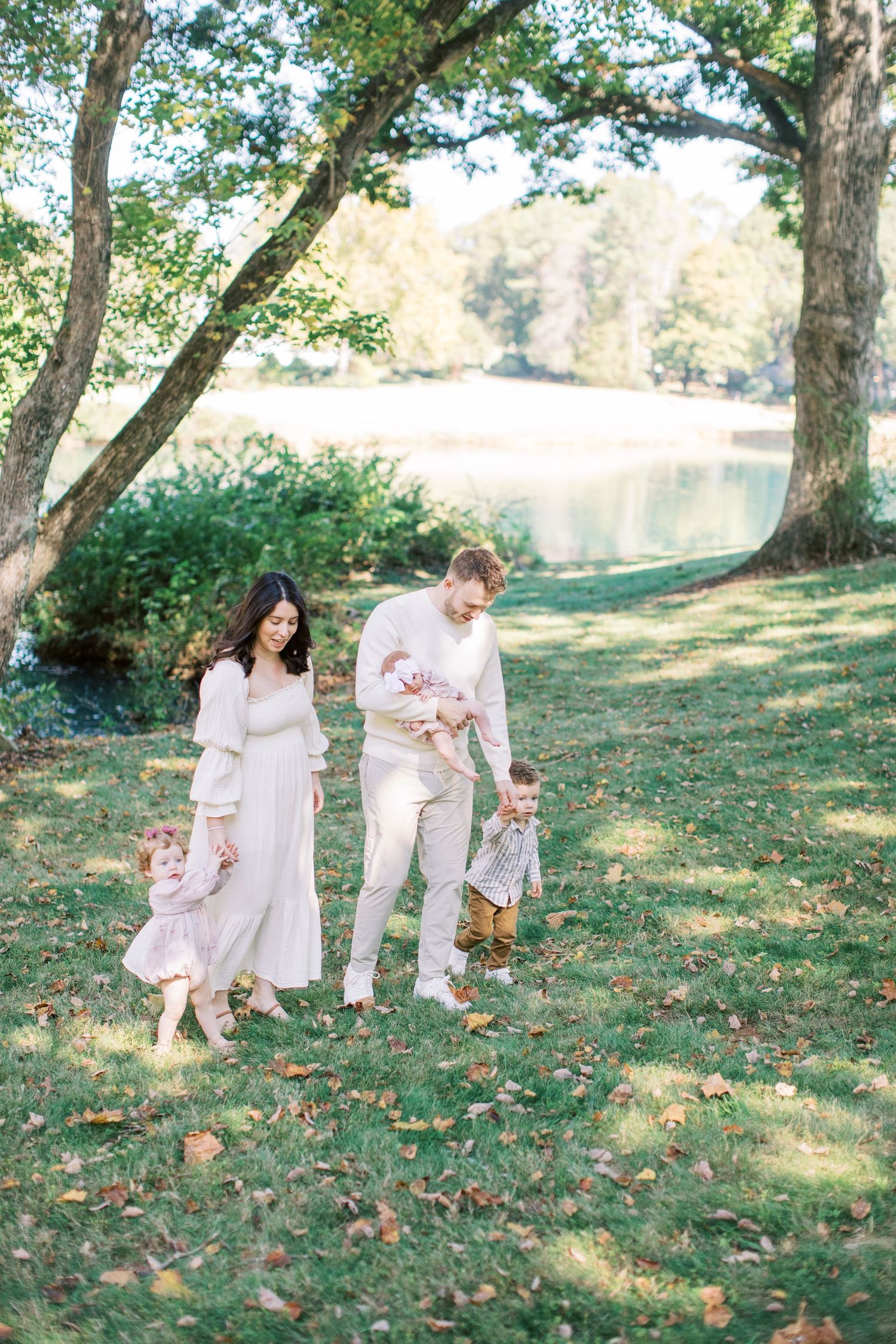Family of four walking outdoors near a pond, parents holding children's hands on green grass under large trees.