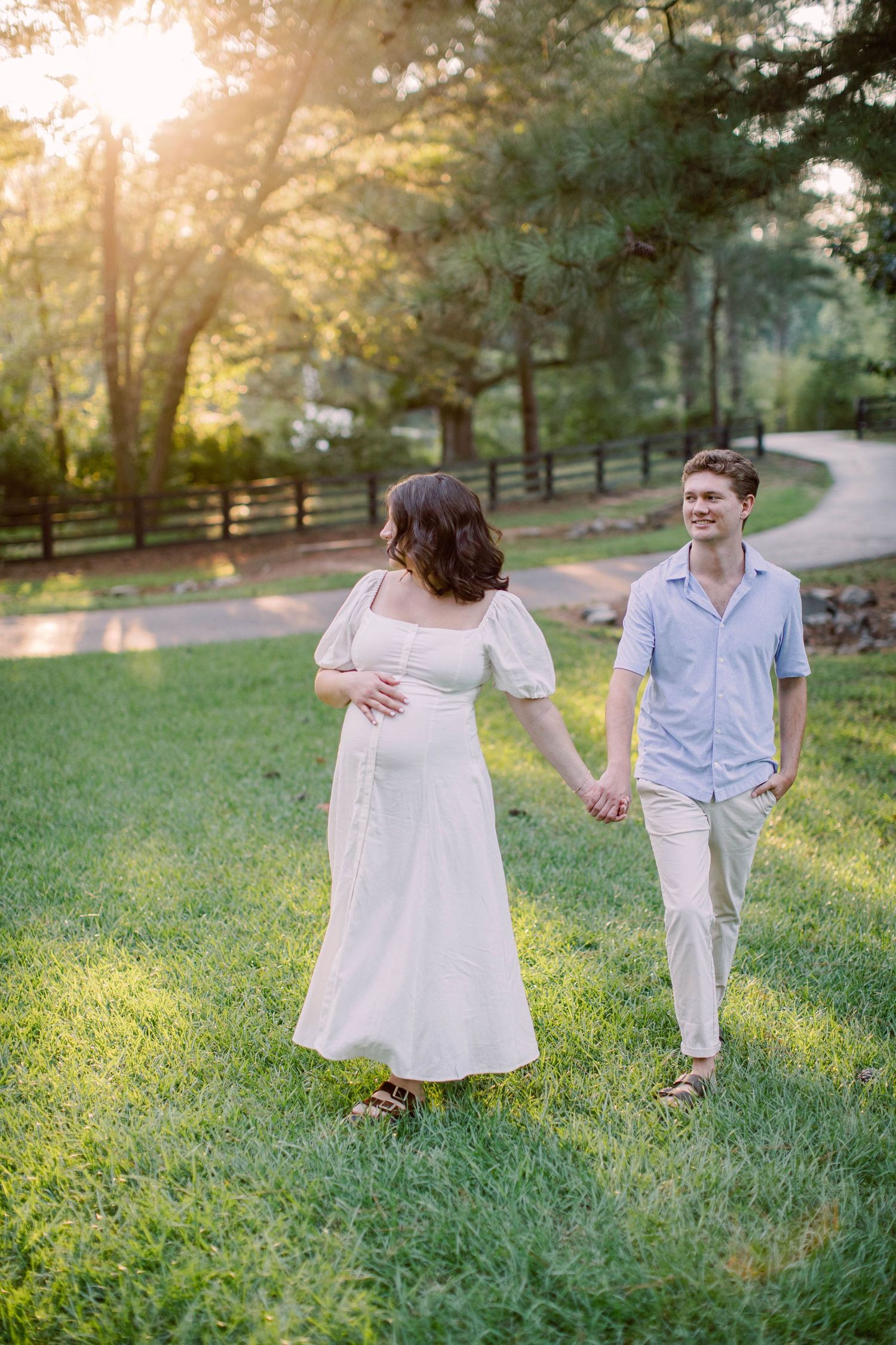Pregnant woman in white dress holds hands with man in blue shirt, walking on green grass at golden hour.