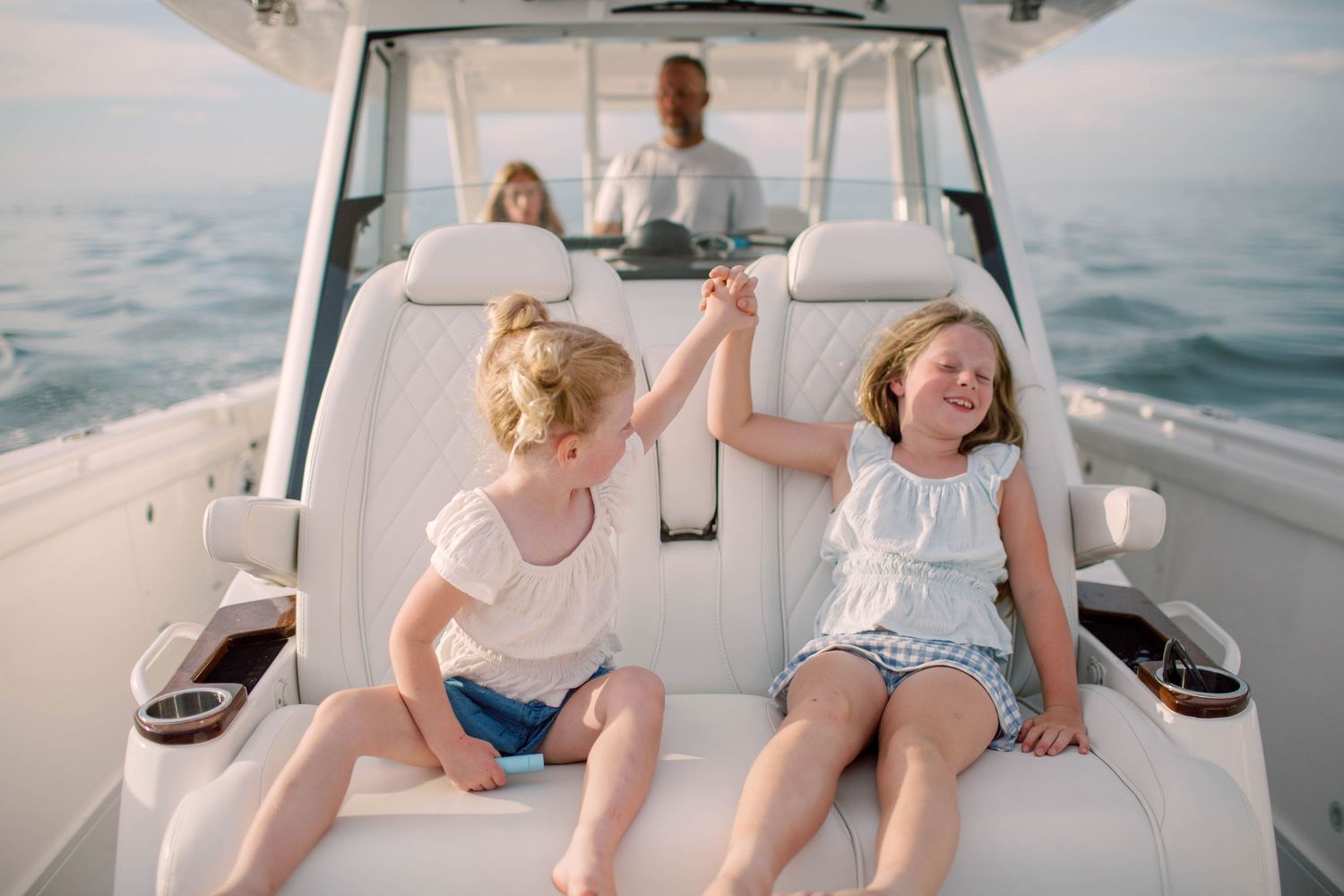 Two young girls in white tops high-five on a white boat deck while adults navigate on calm ocean waters.