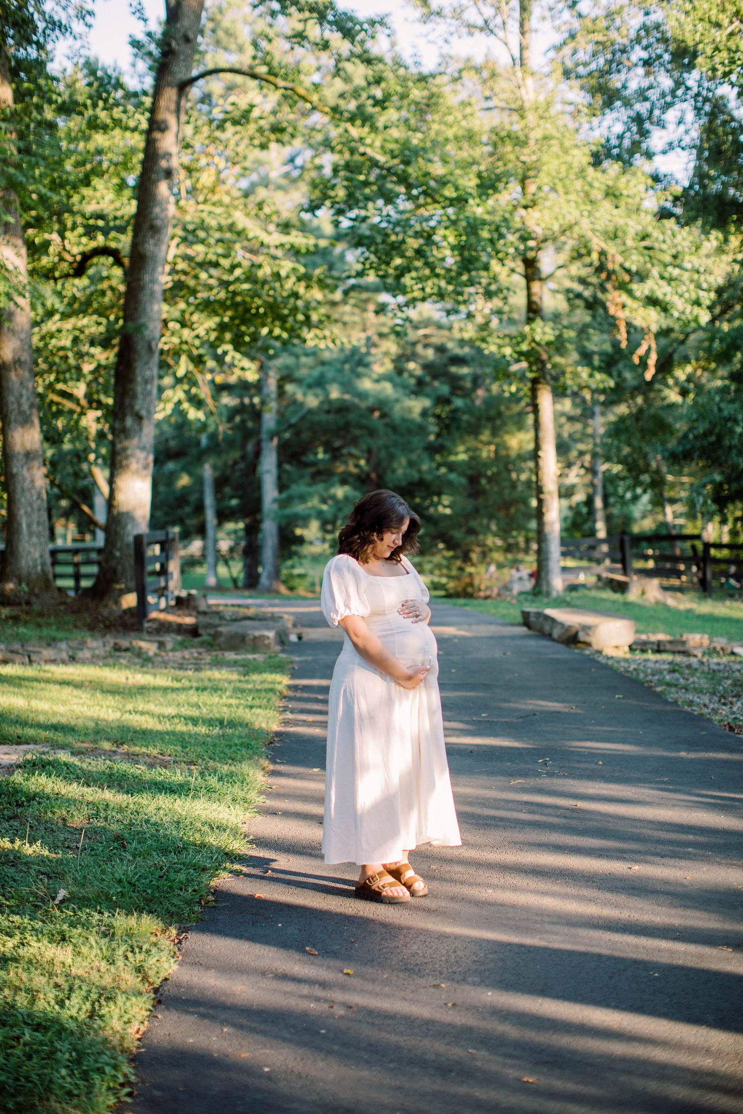 Pregnant woman in a white dress cradling her baby bump while standing on a tree-lined path in soft morning light.