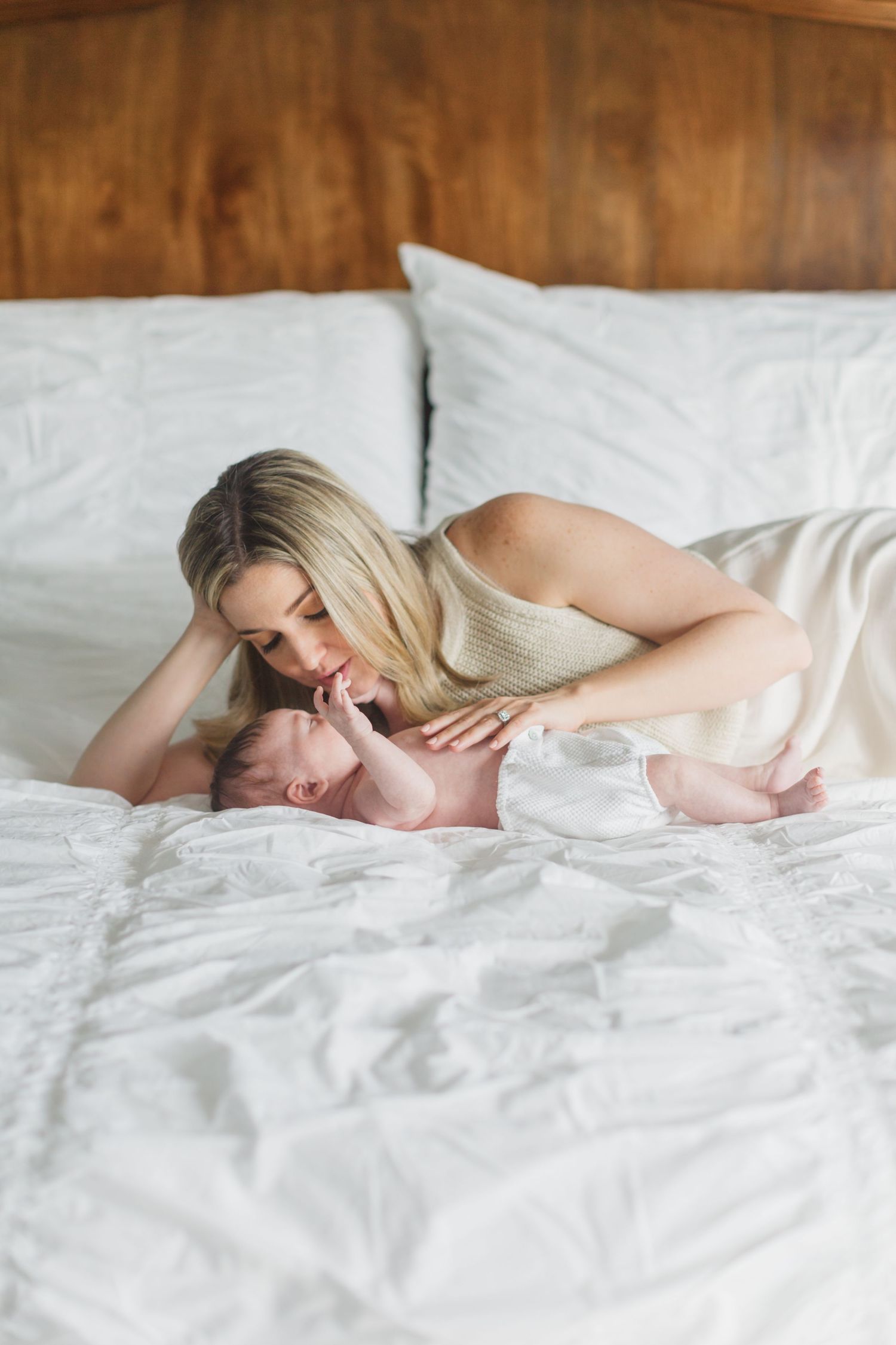 Blonde mother leaning over her newborn baby on a white bed with wooden headboard.