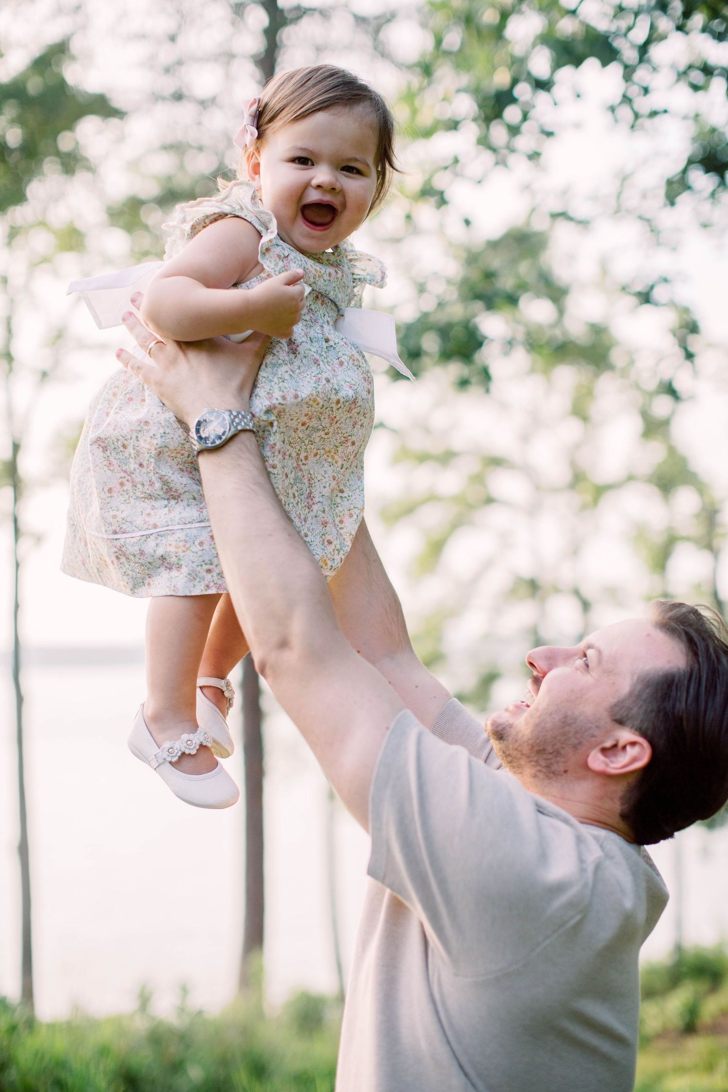 Father joyfully lifts smiling baby girl in floral dress overhead outdoors near lake surrounded by trees.