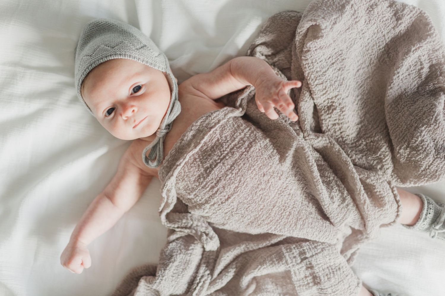 Newborn baby in gray bonnet wrapped in beige muslin swaddle blanket lying on white bedding.