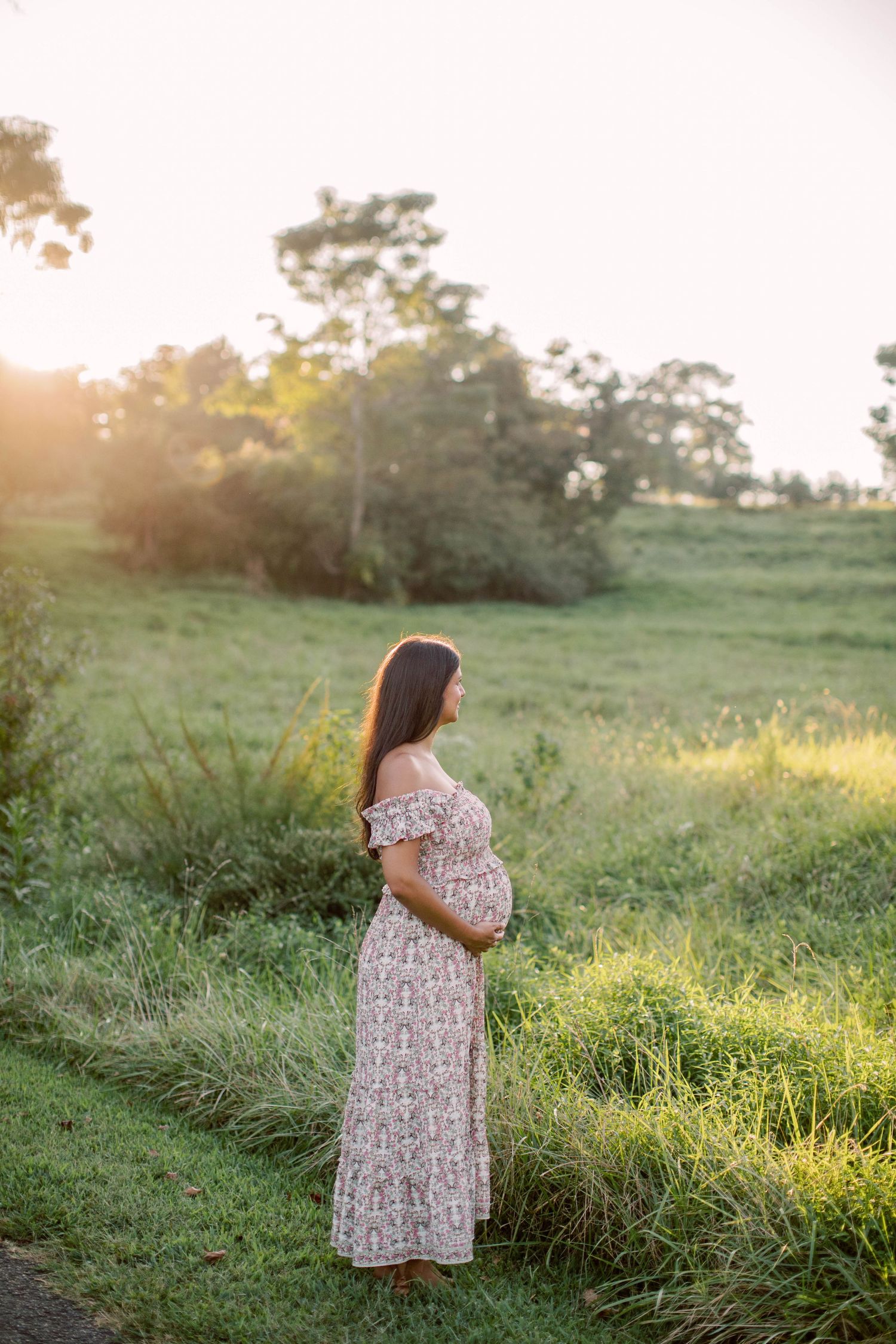 Pregnant woman in floral off-shoulder maxi dress standing in sunlit green field during golden hour maternity photoshoot.