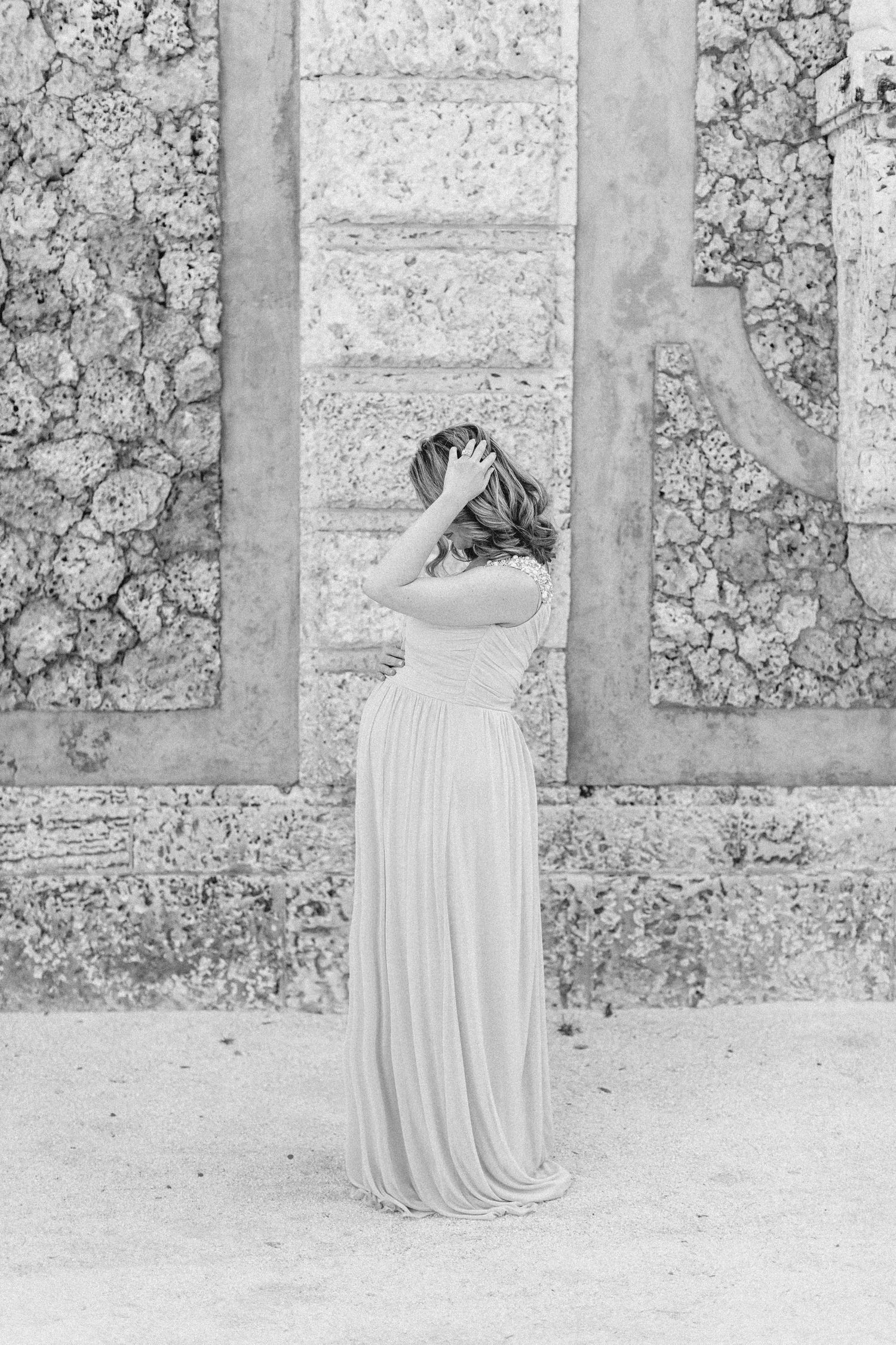 Woman in elegant white gown poses gracefully against a textured stone wall in black and white photography.