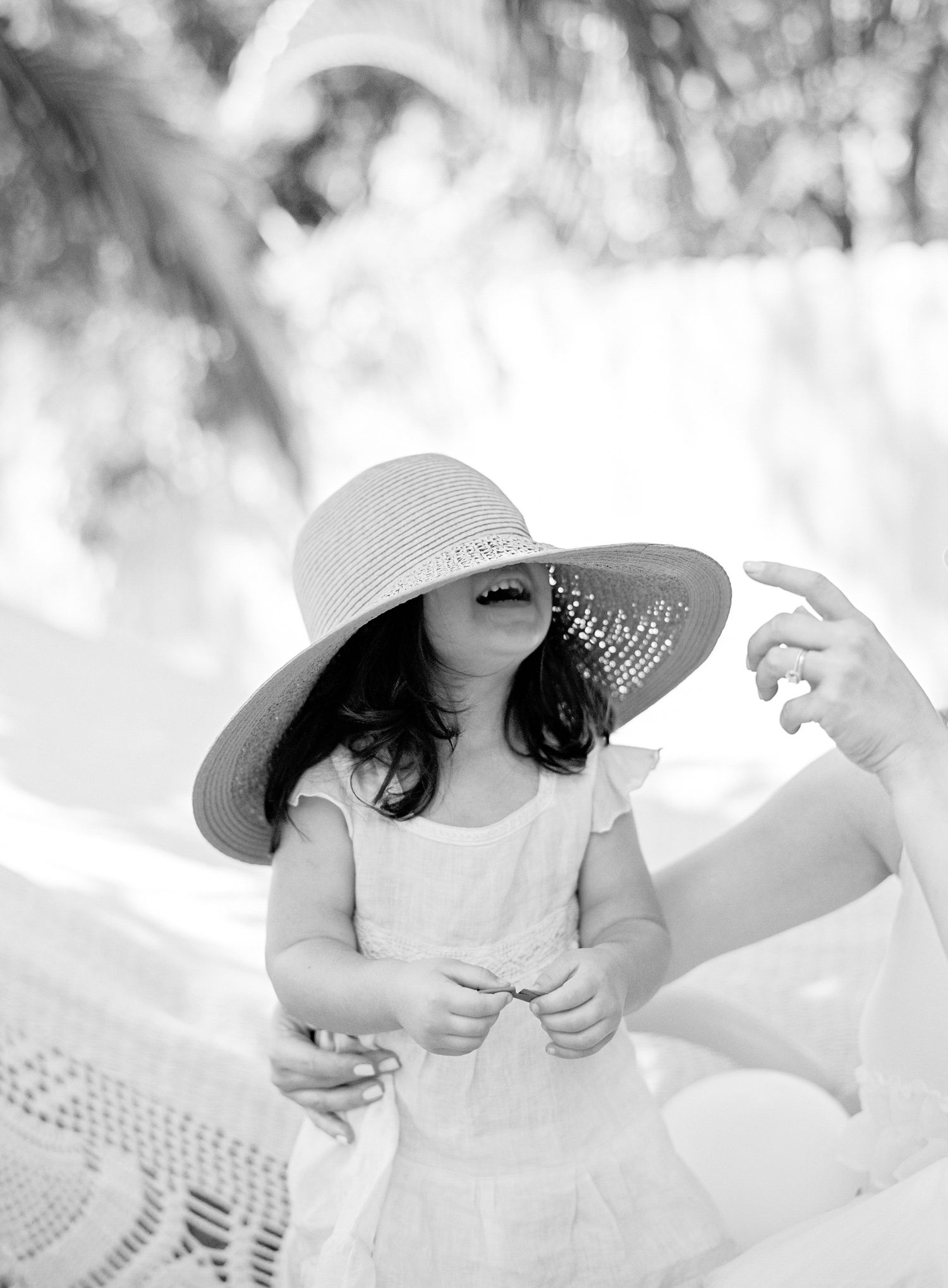 Black and white photo of a laughing toddler girl wearing a large sun hat on a sunny beach.