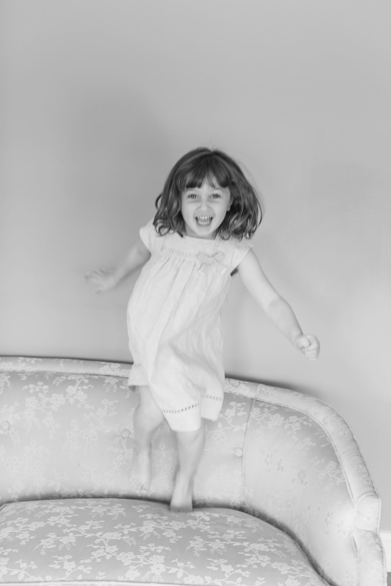 Black and white photo of a joyful young girl in a white dress jumping on a floral sofa, laughing with arms outstretched.