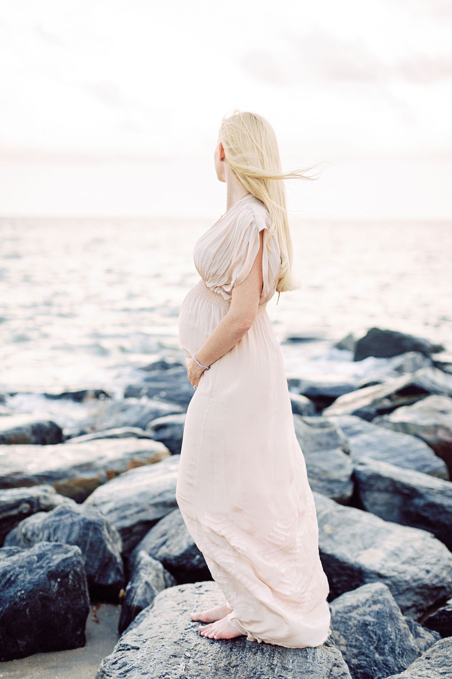 Pregnant woman in a flowing blush gown standing on rocks by the ocean at sunset.