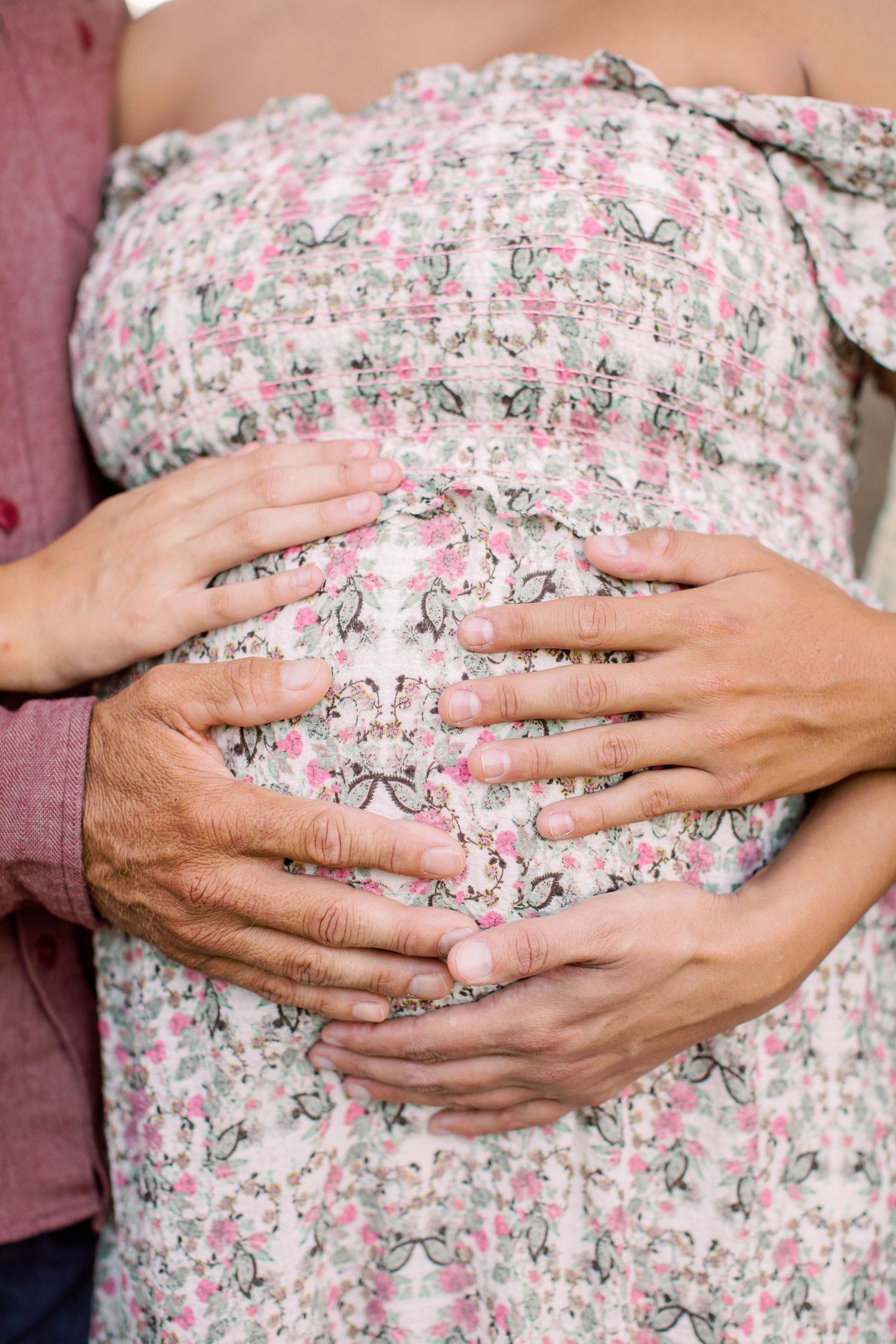 Couple cradling pregnant belly, hands forming heart shape over floral maternity dress.