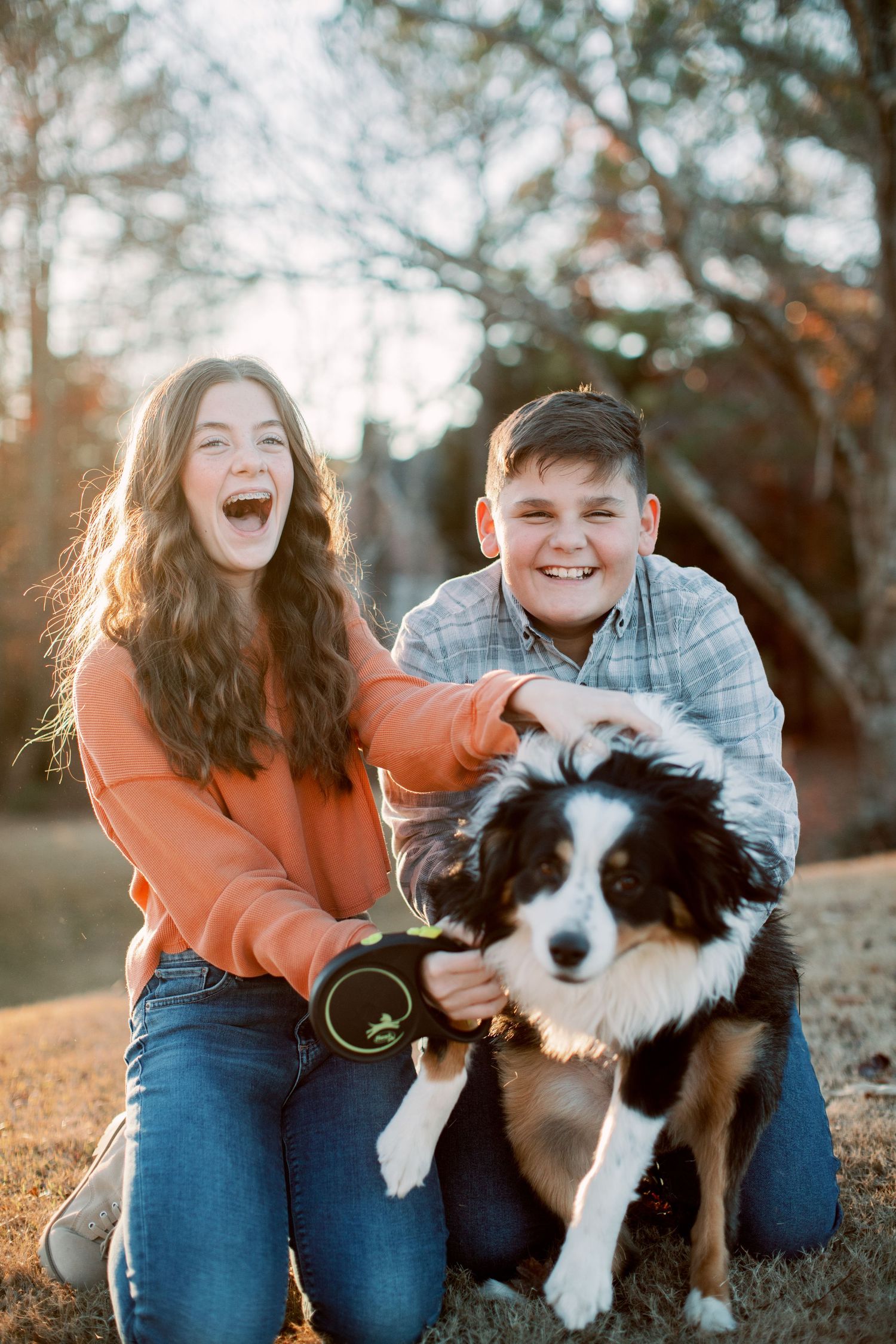 Two laughing kids petting a black and white Border Collie puppy outdoors in golden hour light.