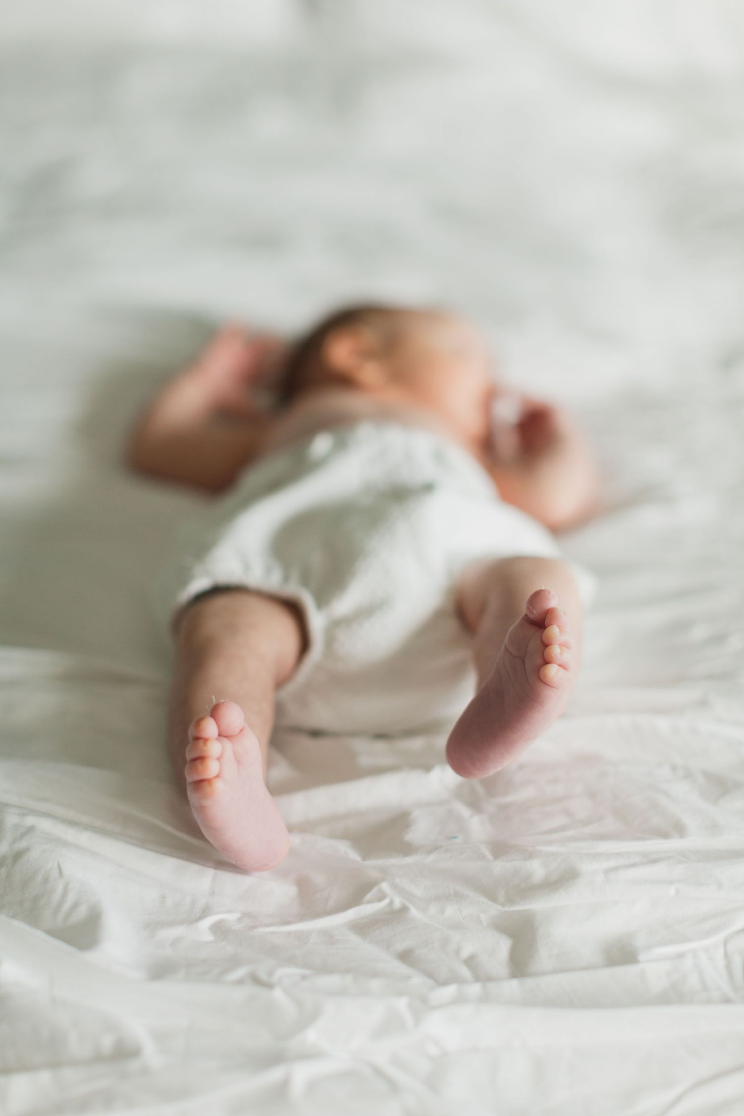 Newborn baby lying on white bedding with tiny feet in focus, wearing a white onesie in a soft lifestyle photo.
