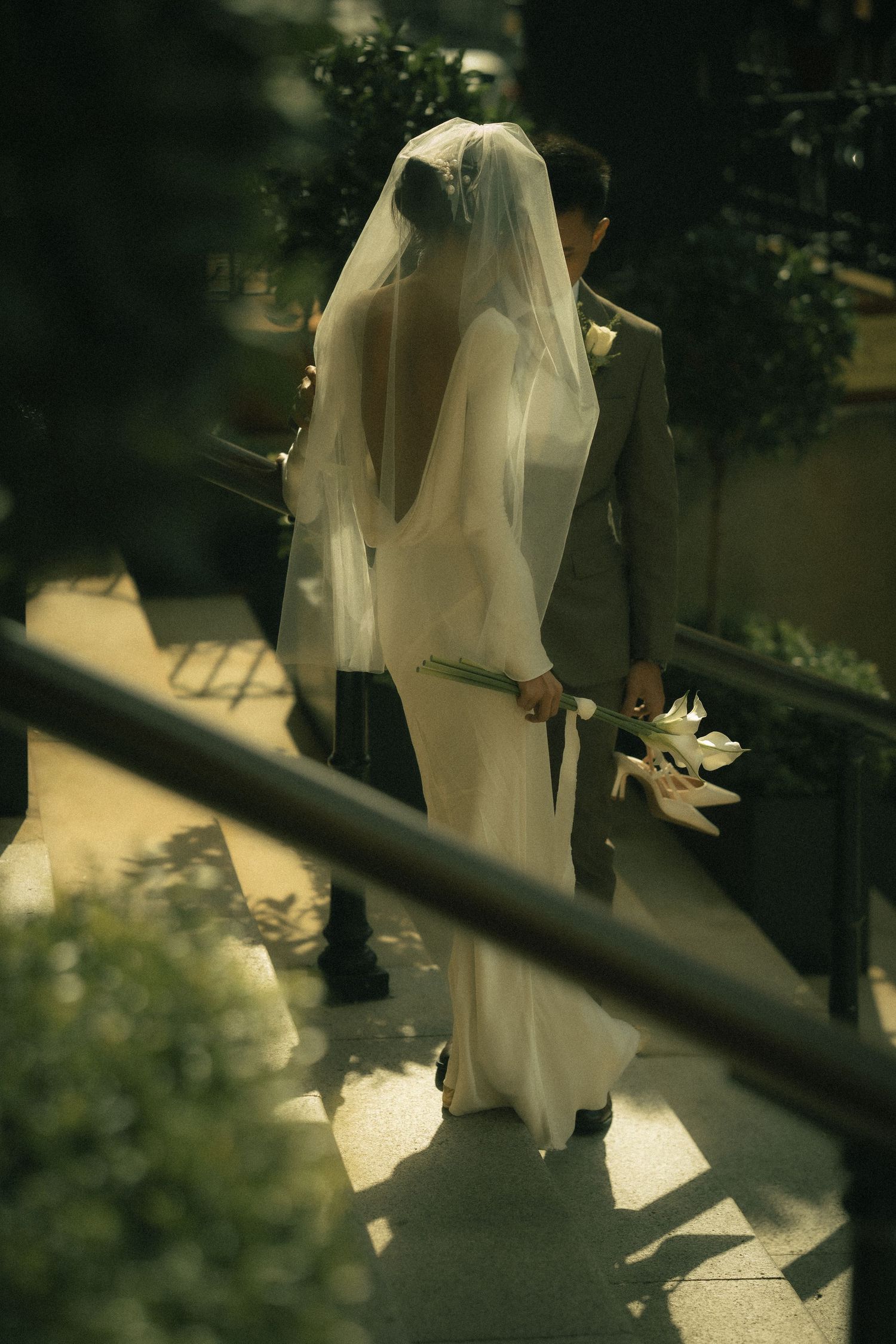 Atmospheric portrait of a bride and groom standing together on steps with their backs to the camera, surrounded by soft natural light.