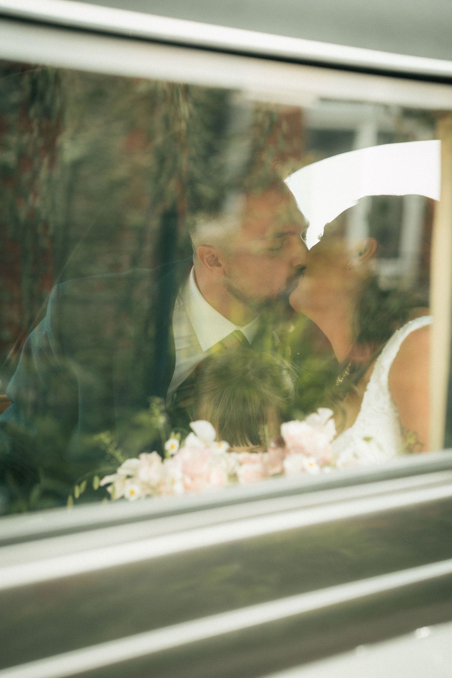 Bride and groom kissing behind the glass window of a wedding car, in gentle vintage style lighting.