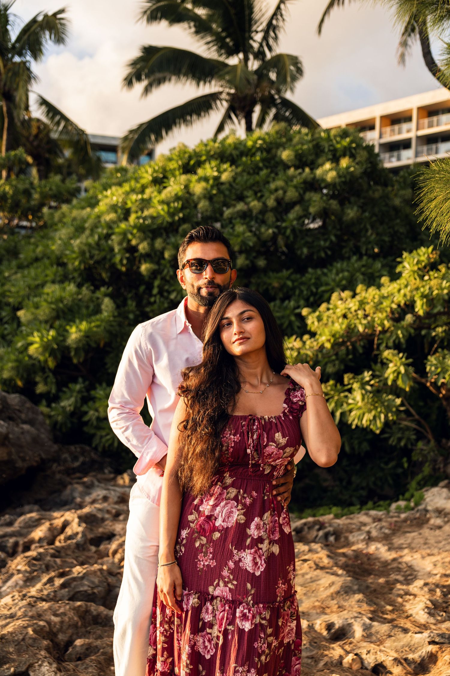 South Asian couple posing on rocky Hawaiian beach, woman in floral dress, man in pink shirt.