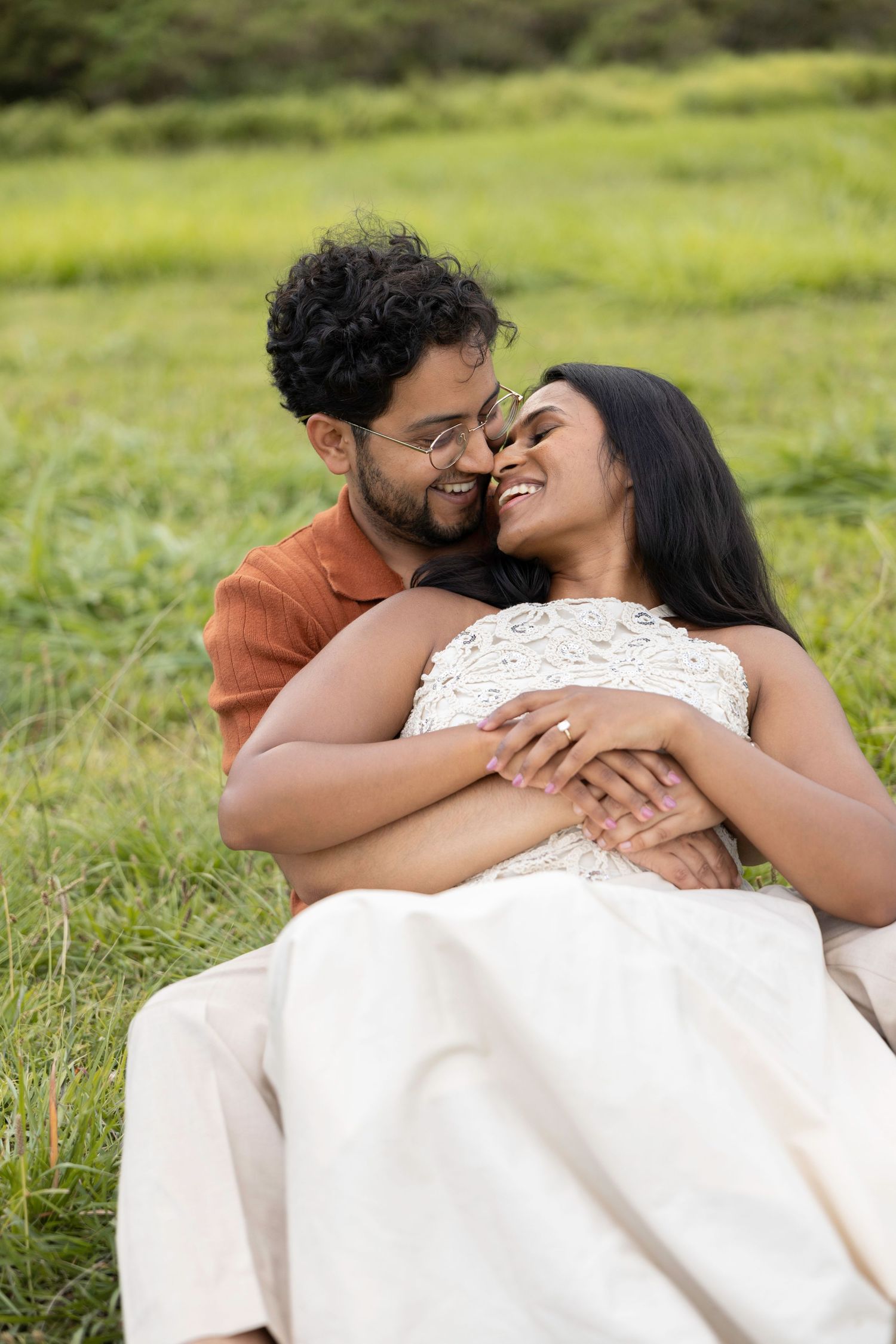 Couple embracing in a green meadow, woman in white dress smiling as man holds her close.