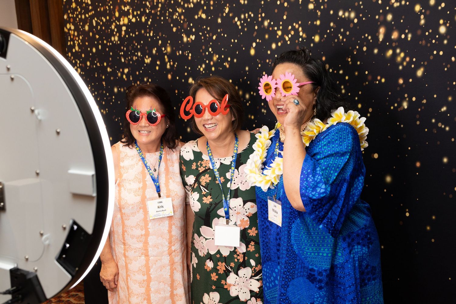Three women wearing fun novelty sunglasses pose together at a photo booth with a sparkly black backdrop.