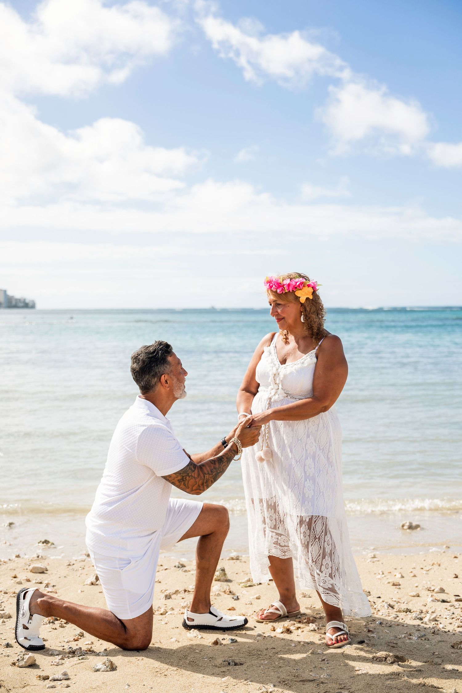 Man proposing on one knee to woman in white dress and floral crown on a sunny tropical beach.