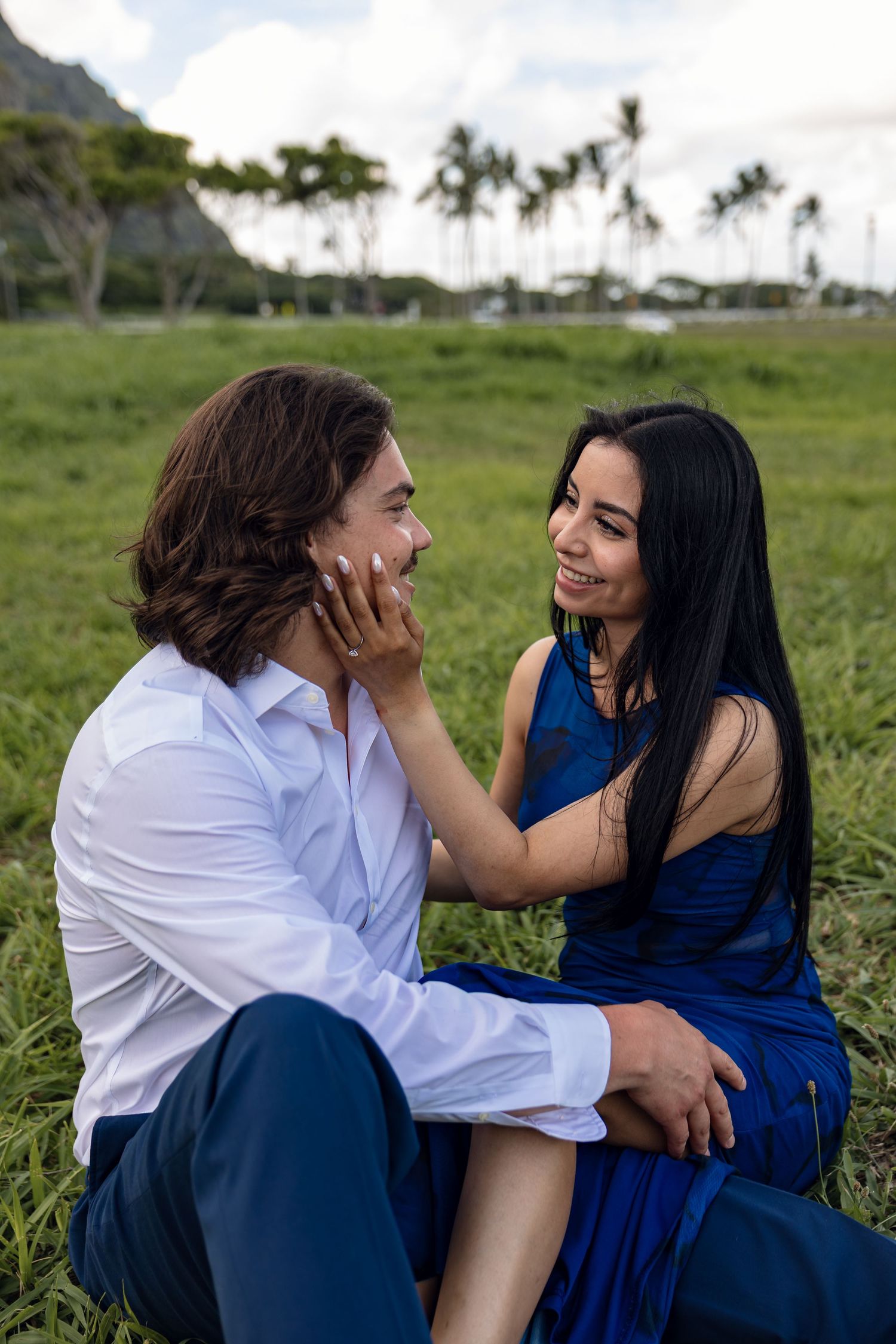 Smiling couple sitting on grass in Hawaii, woman in blue dress tenderly touching mans face.