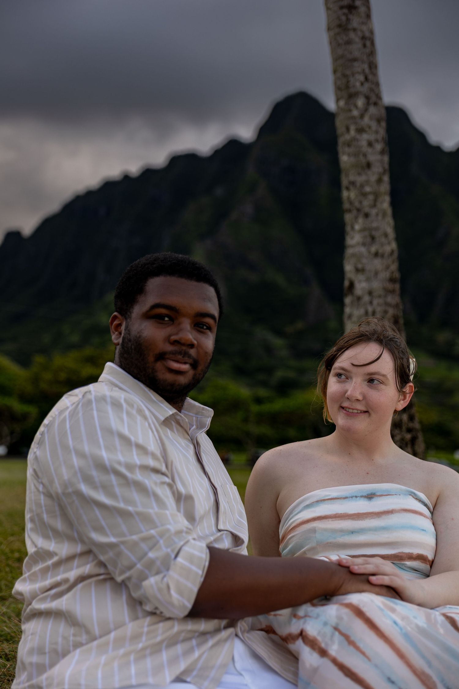 A couple sits together on grass with stunning dark mountain scenery behind them, the man in a striped shirt holding her close.