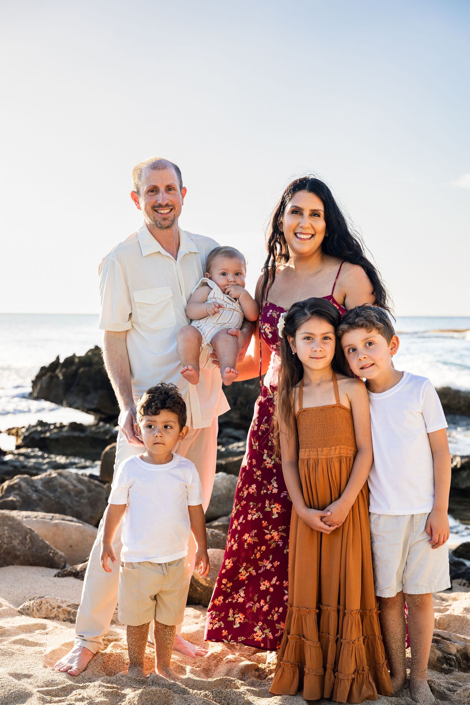 Family of six posing on a rocky beach at sunset, parents with four children smiling in coordinated neutral and earth-tone outfits.
