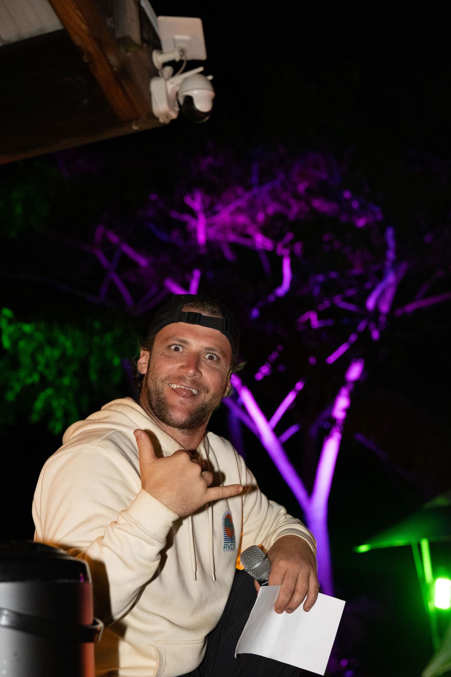 Man in beige hoodie and cap holds microphone at night event with purple and green lit trees in background.