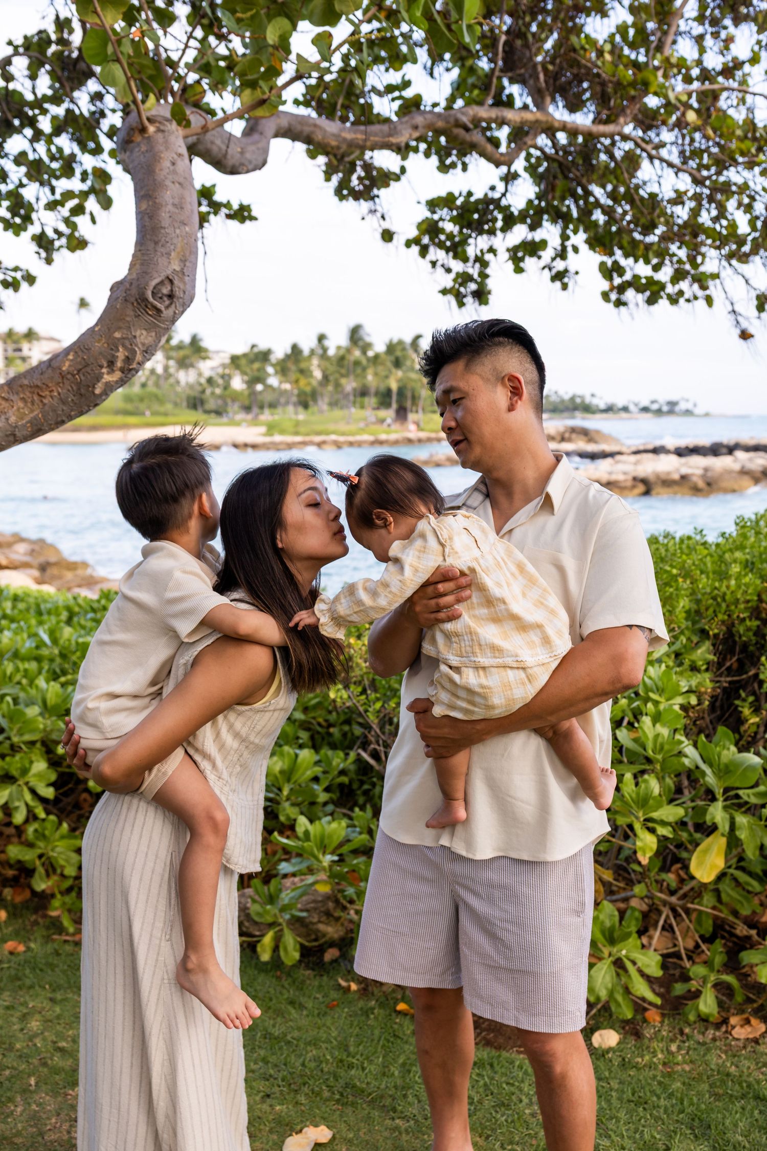 Young Asian family with two toddlers sharing a tender moment under a tree by the coast.