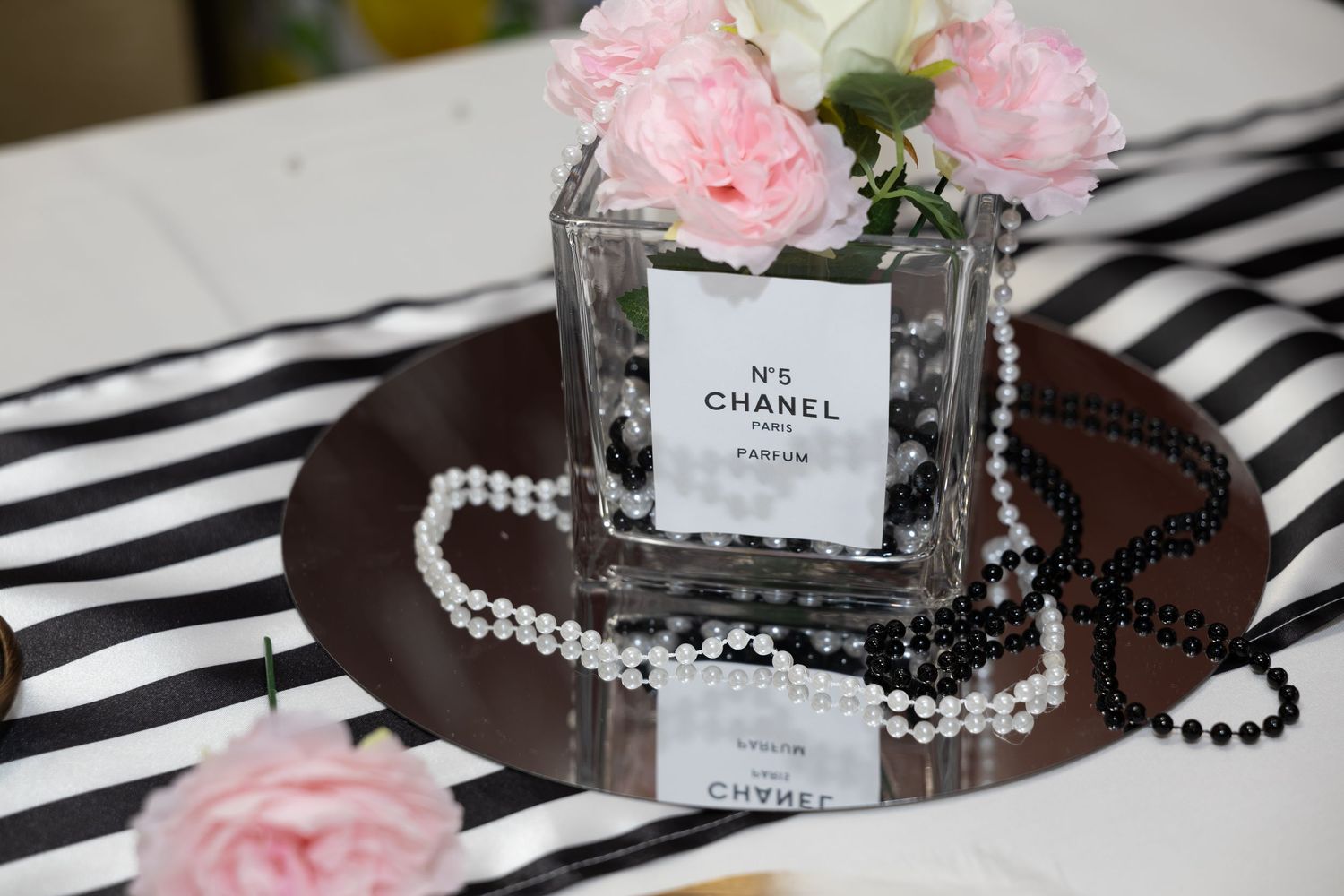 Chanel No.5 perfume bottle vase with pearl necklace on striped mirror tray surrounded by pink roses.