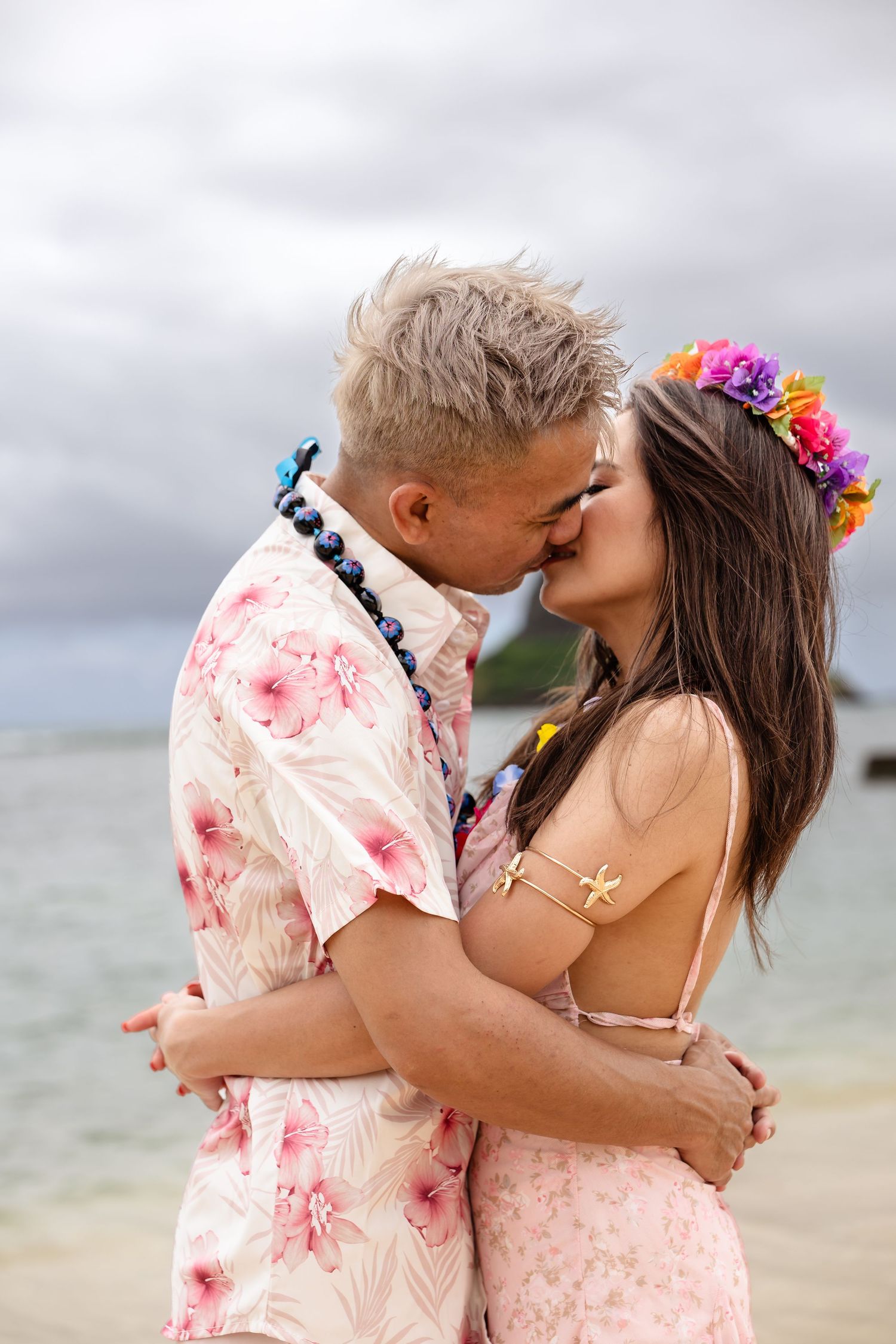 Couple sharing a kiss on the beach, woman wearing vibrant floral crown and starfish bikini top.