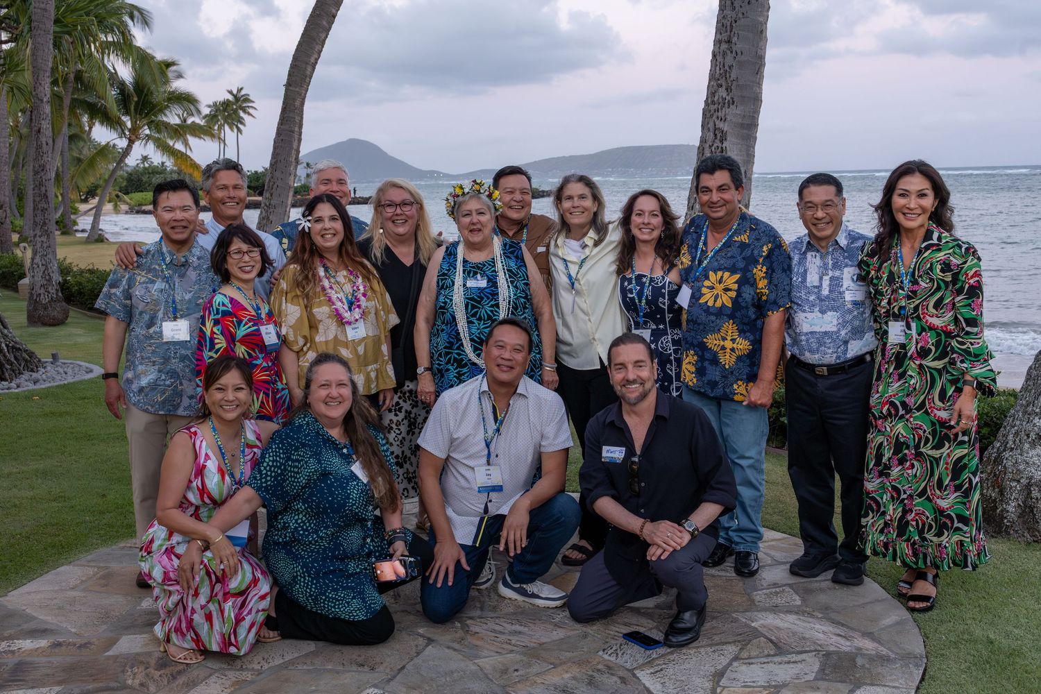 Group of professionals posing outdoors near ocean with palm trees in Hawaii, wearing colorful tropical attire at a networking event.
