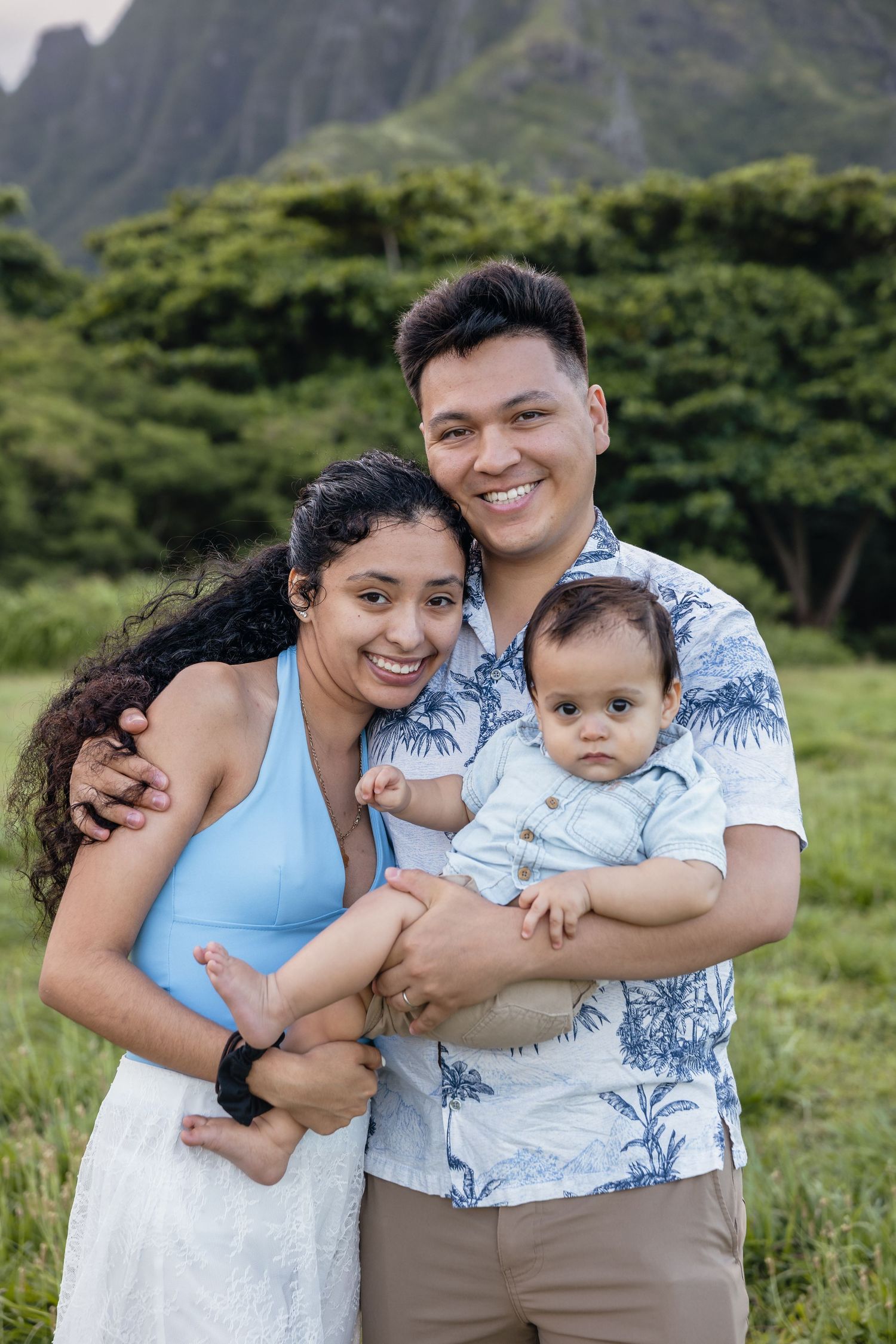 Happy young family with baby posing outdoors in Hawaii with lush green mountains in the background.